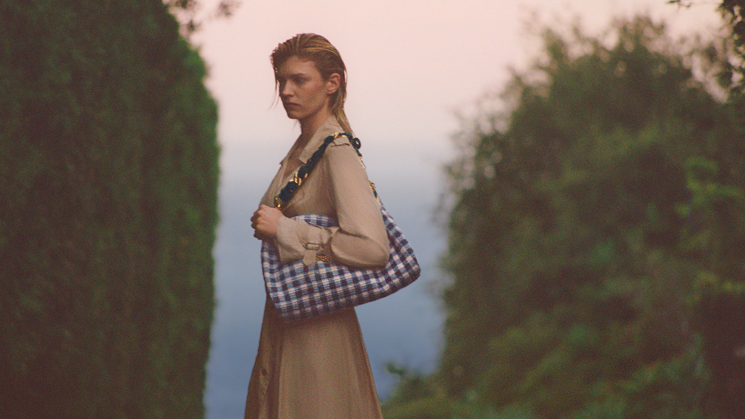 Model walking between tall hedges at sunset carrying a blue gingham shoulder bag and wearing a beige trench coat.			
