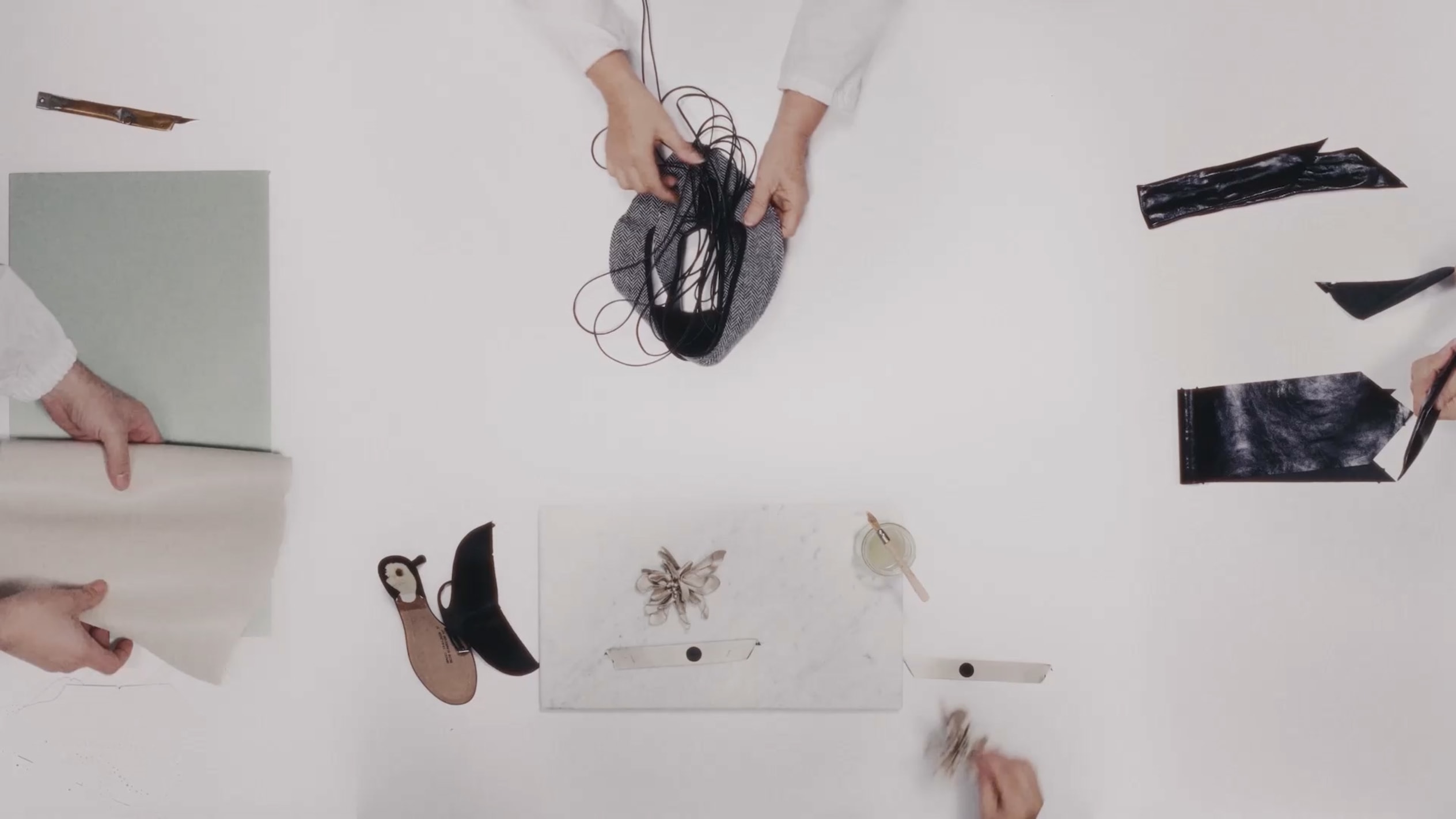 Overhead view of hands arranging design materials, sketches and accessories on a white worktable.