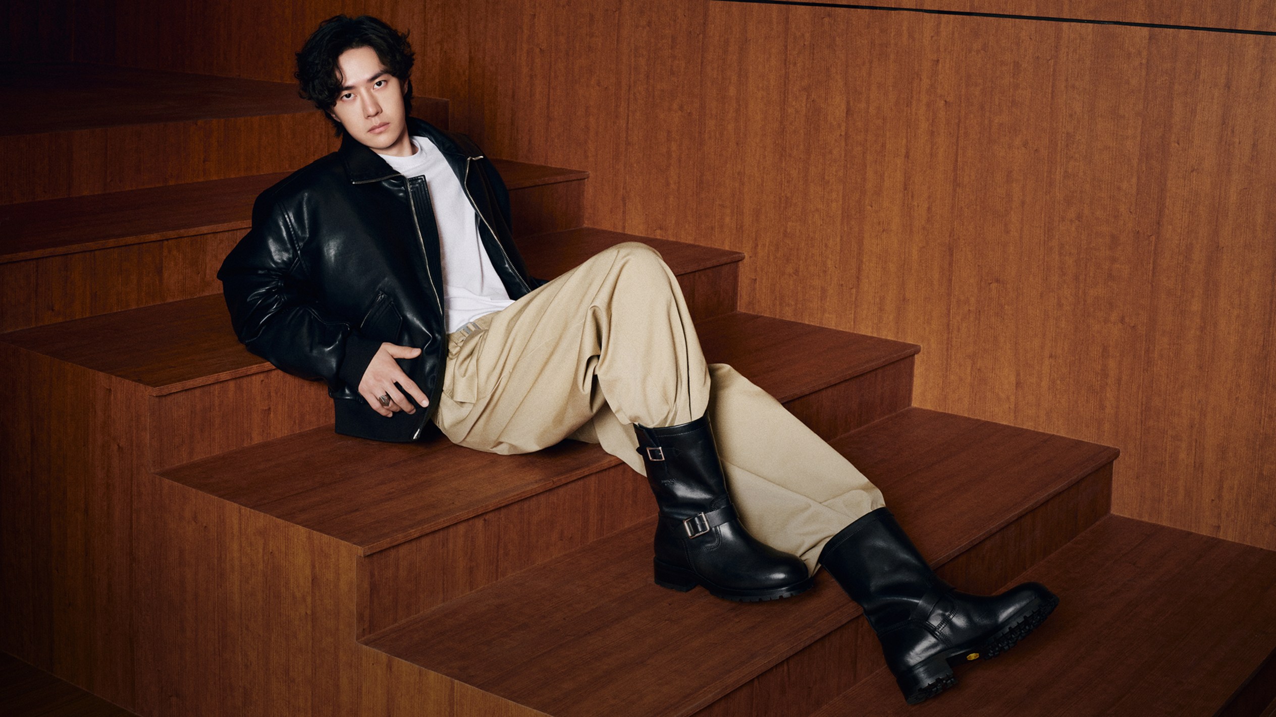 Wang Yibo seated on wooden steps wearing a black leather jacket, beige trousers and black leather biker boots.