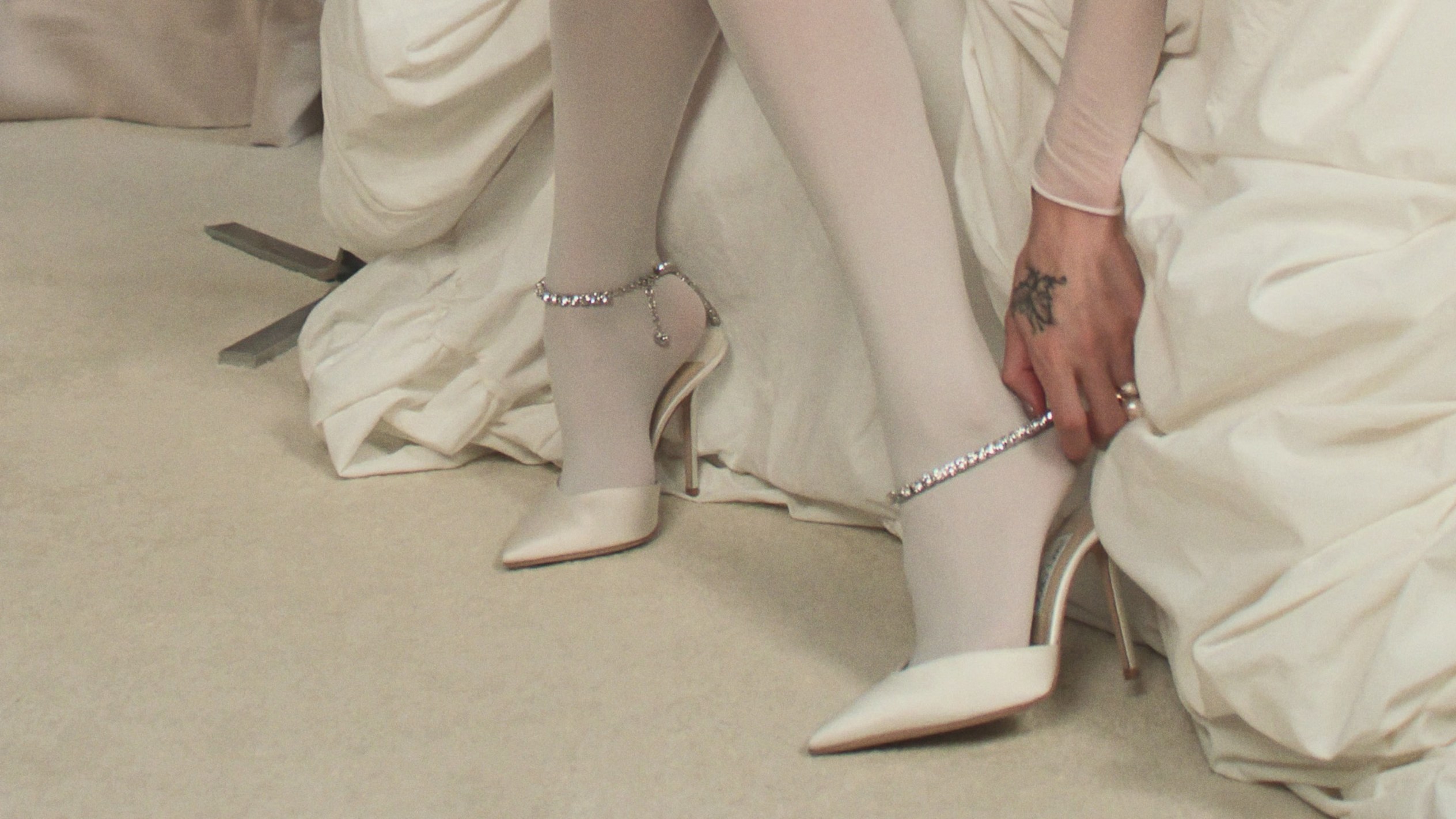  Model wearing ivory bridal heels with pearl detail, holding white flowers indoors