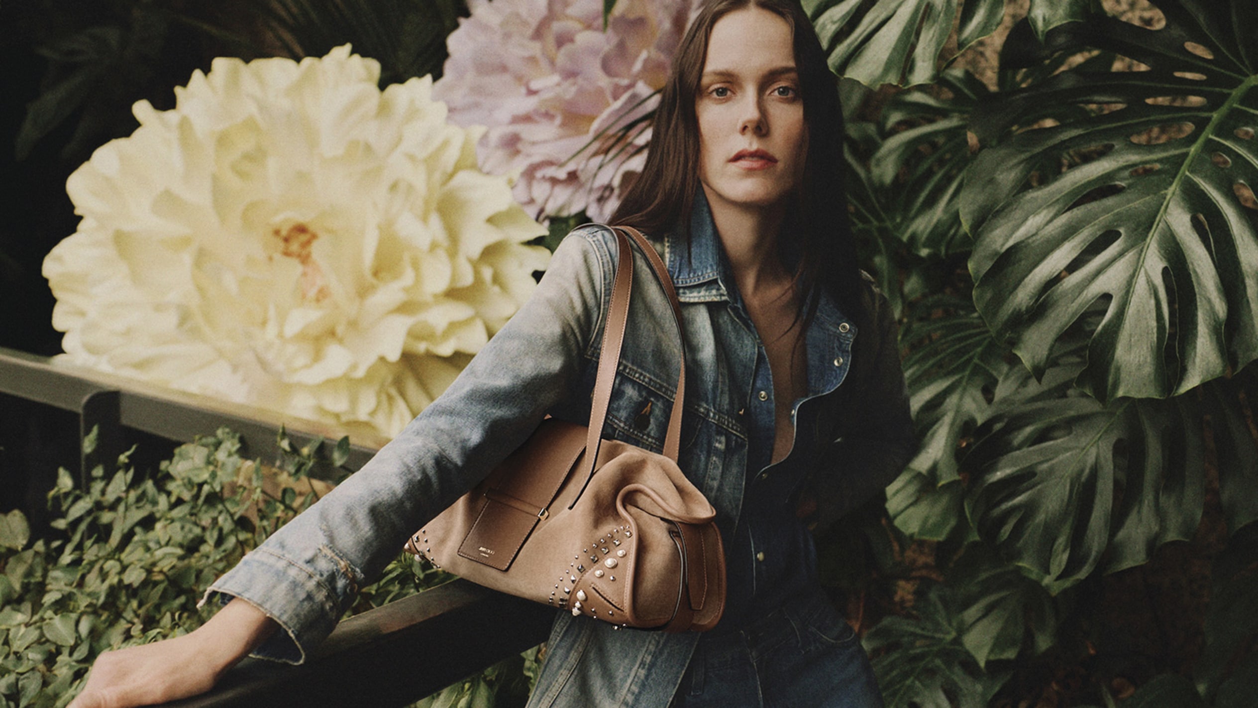 Model wearing a denim jacket and jeans, carrying a brown leather shoulder bag with stud detailing, posed in front of large floral installations and green foliage.