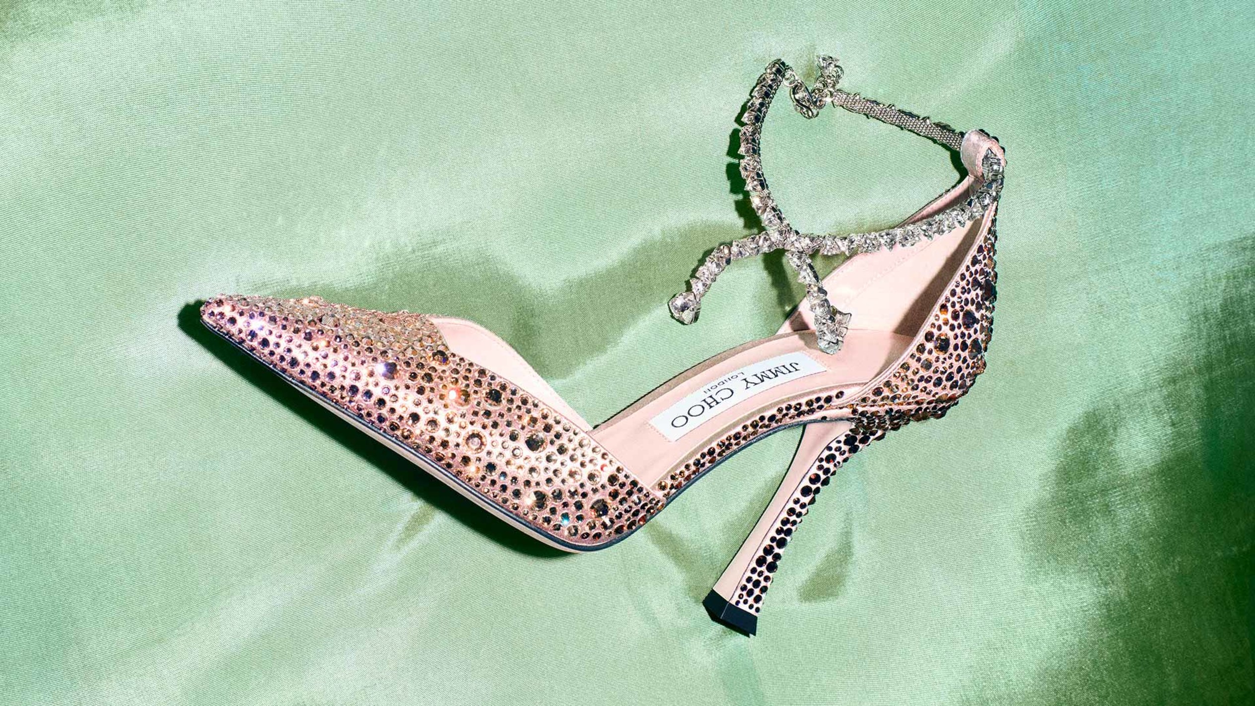 Close-up of a sparkling pointed-toe pump embellished with crystals and a delicate ankle strap, set against a soft green background.