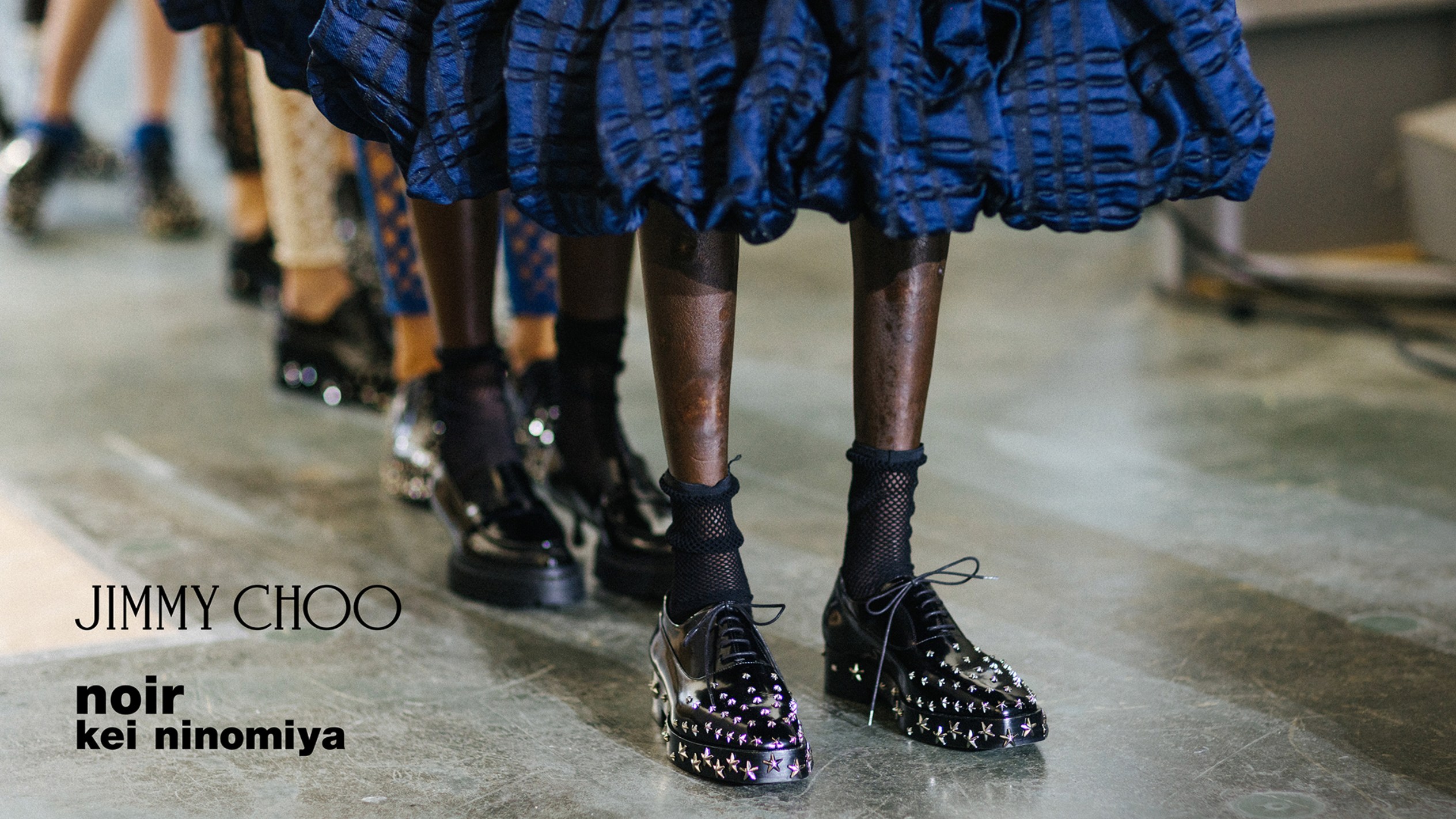 Multiple models standing in a row wearing black leather studded lace-up shoes with sheer socks, styled backstage.