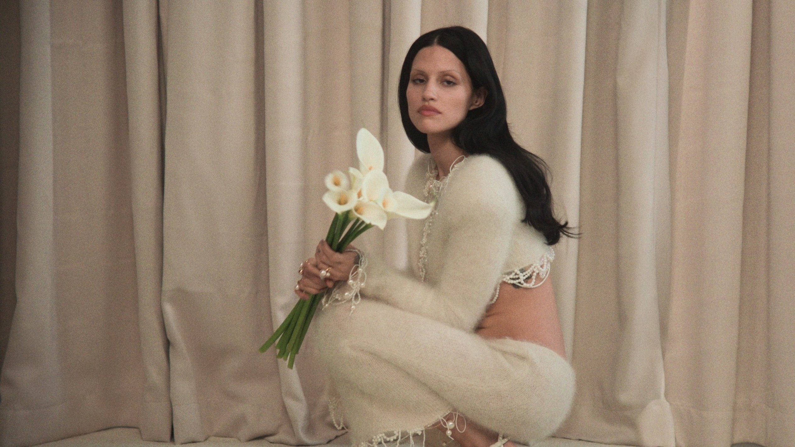 Model wearing ivory bridal heels with pearl detail, holding white flowers indoors./ Ivory open-toe heels with pearl bow embellishment and ankle strap, shown close up.