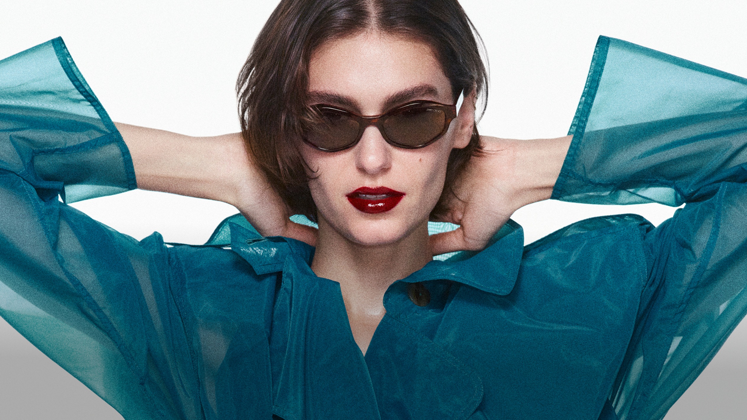 Model wearing sunglasses and a sheer turquoise coat, posing with her hands behind her head against a white backdrop.