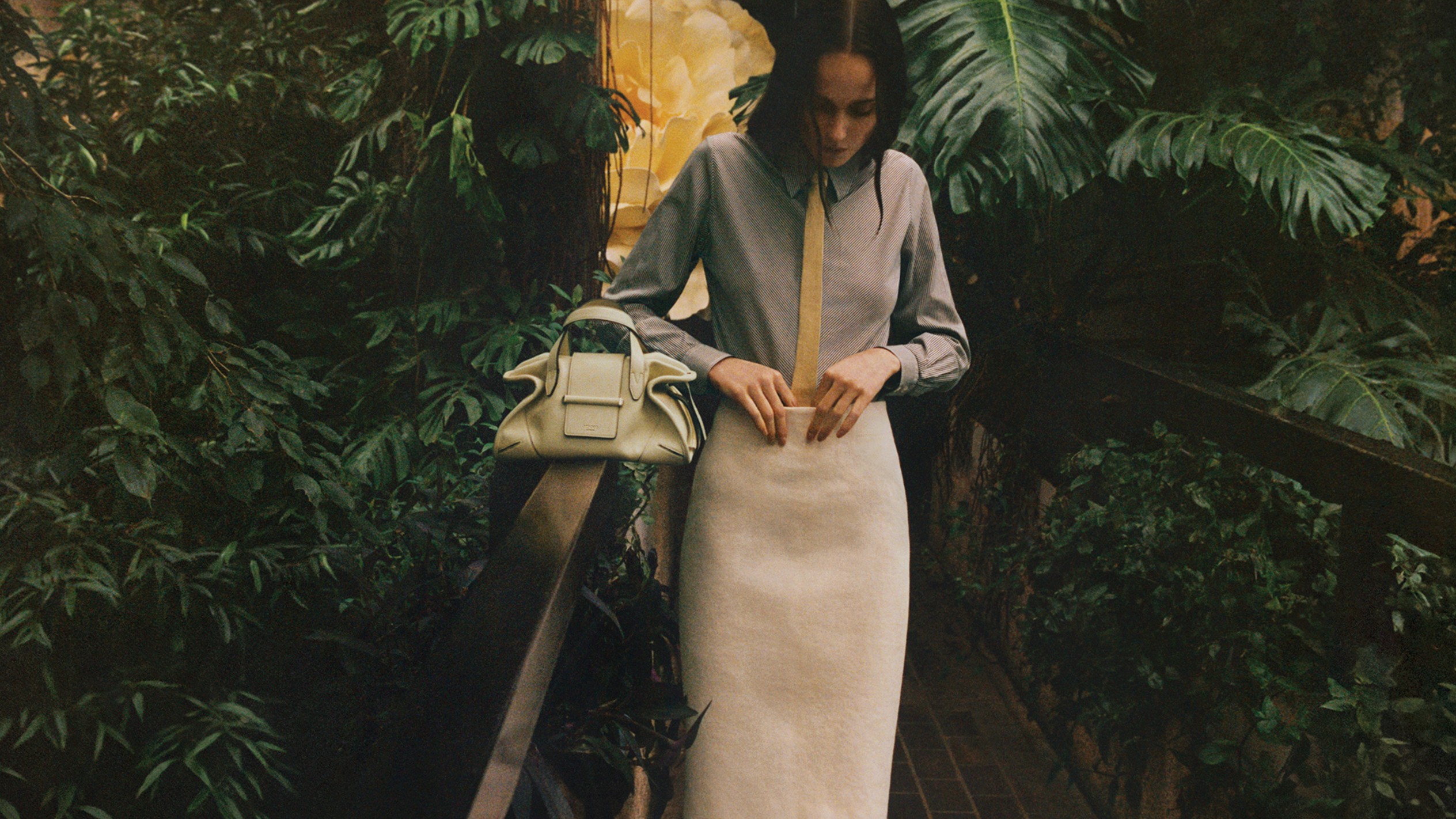 Model wearing a cream skirt and blouse, styled with a light yellow top-handle bag in a lush garden setting.