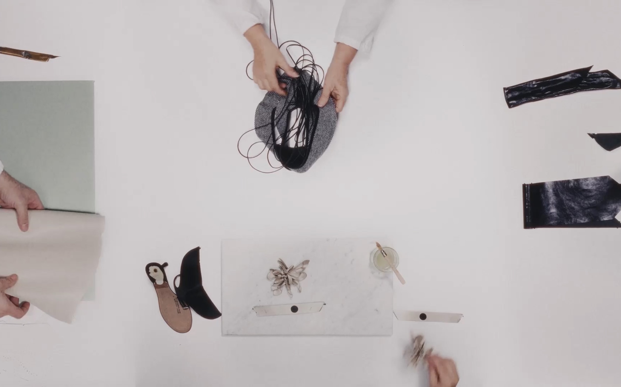 Overhead view of hands arranging design materials, sketches and accessories on a white worktable.
