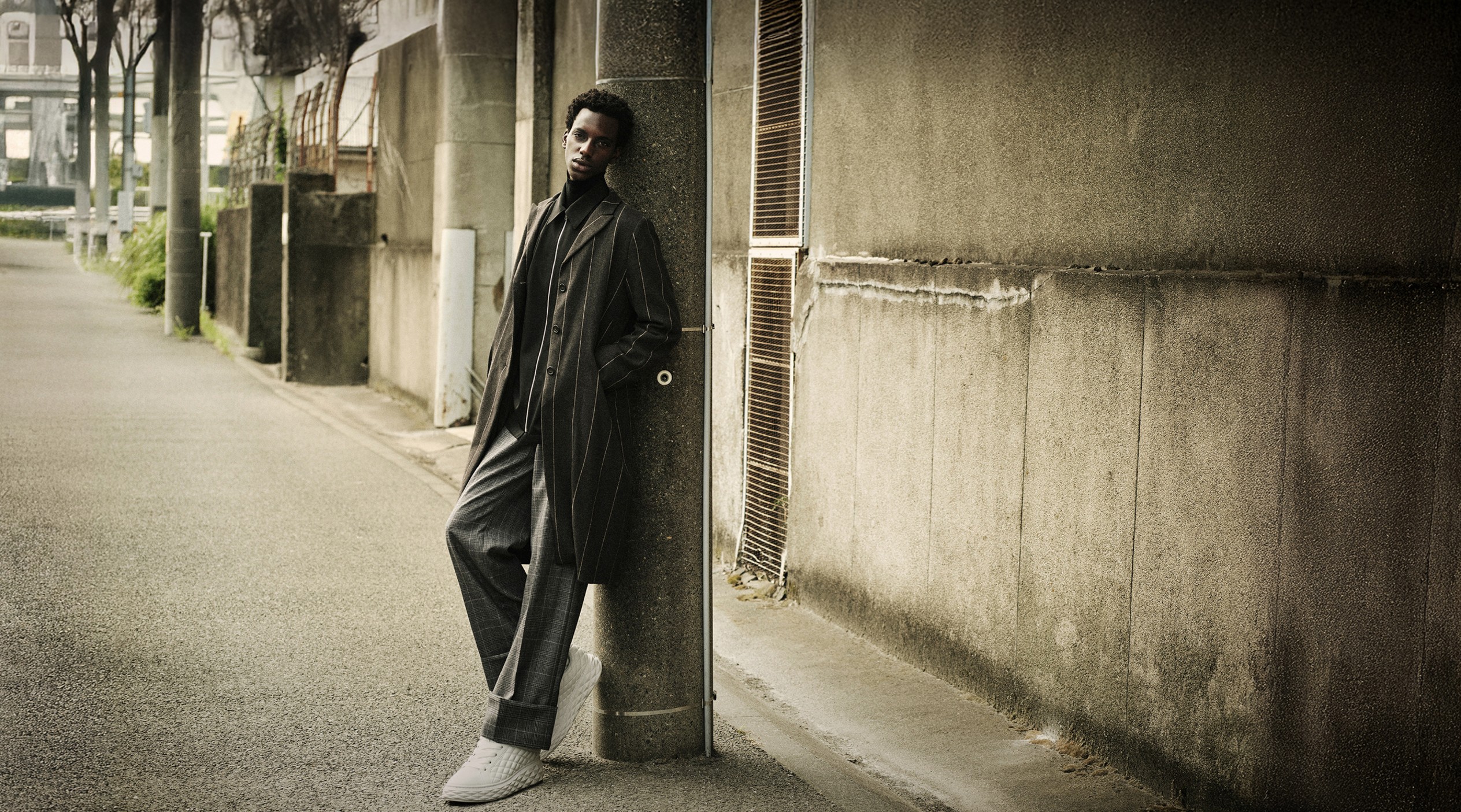  Man leaning against a concrete post in an urban street, wearing a lomg dark coat, striped trousers, and white sneakers.