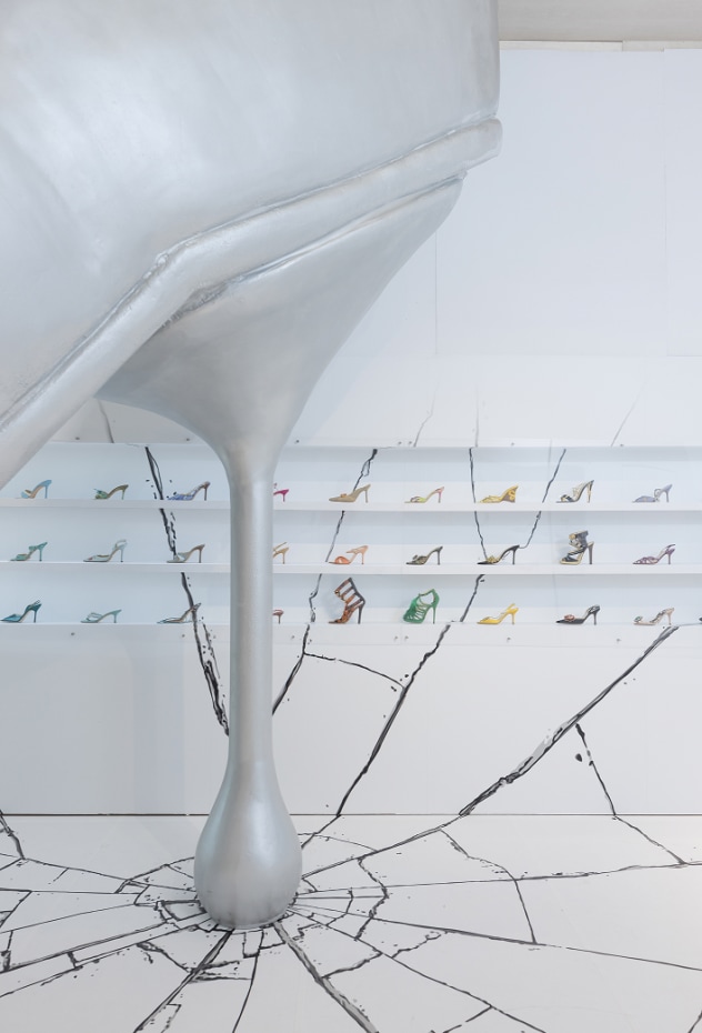 A large silver sculptural high-heel installation emerging through the gallery floor, surrounded by cracked detailing, with shelves of colourful Jimmy Choo shoes displayed along the walls.