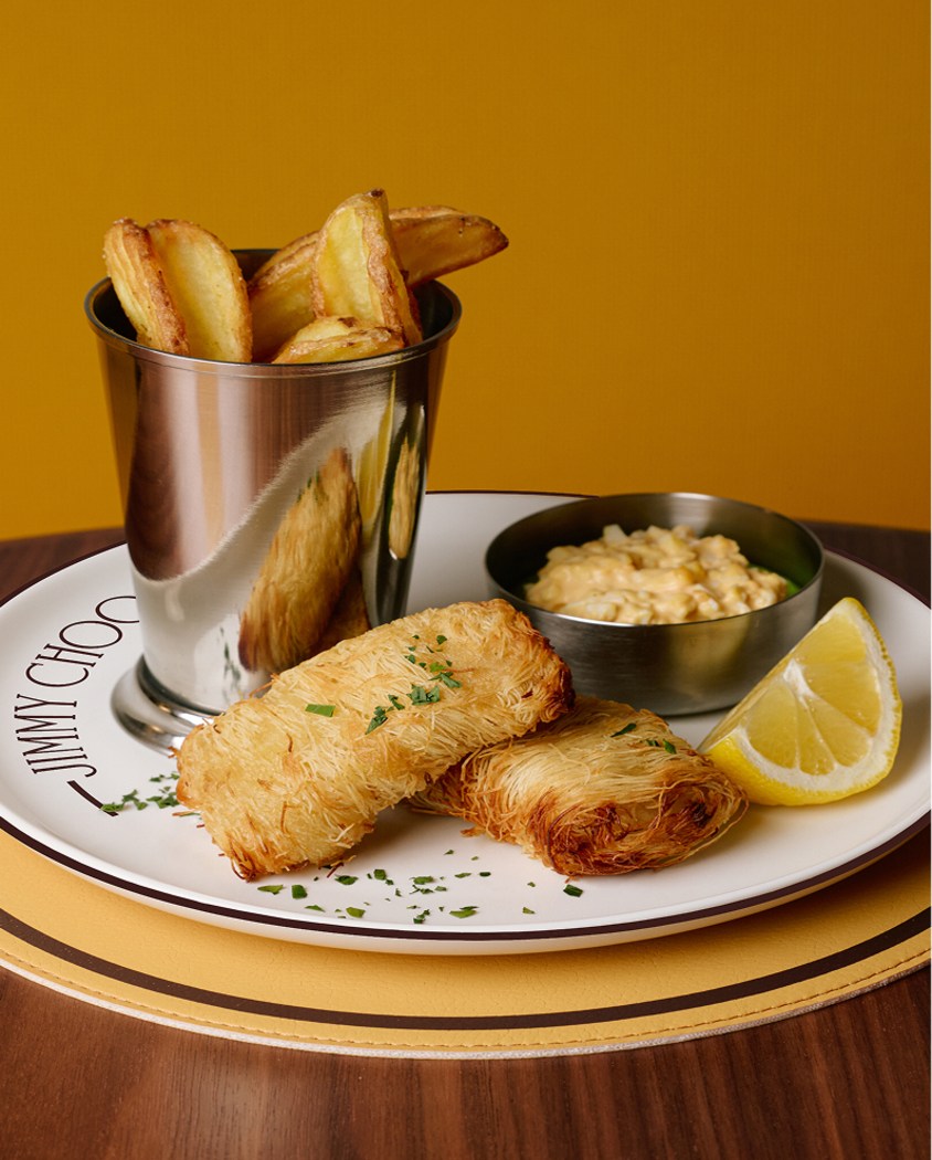 Jimmy Choo ブランドのプレートに盛り付けられた、金属カップ入りのフライドポテトを添えたフライドフィッシュ。