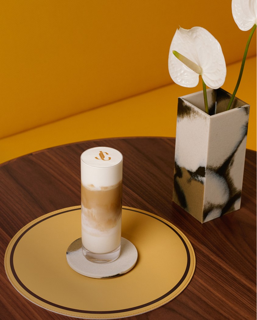  木製テーブルの上で、小さな鉢植えの横に置かれた、ミルクとコーヒーが層になったアイスラテ（黄色い縁のプレートで提供）。