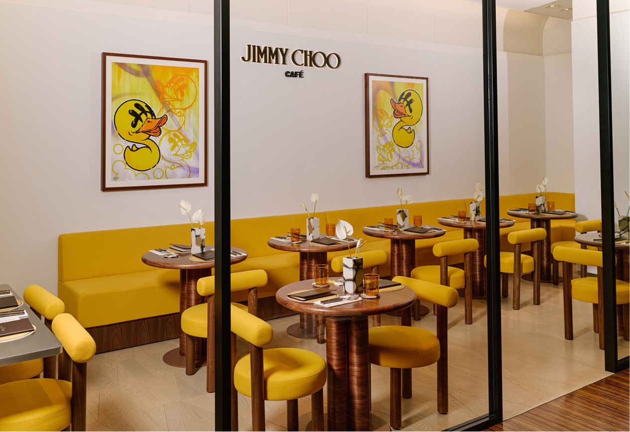 Jimmy Choo Caf&eacute; interior with yellow upholstered chairs and small tables arranged in rows behind a glass partition.