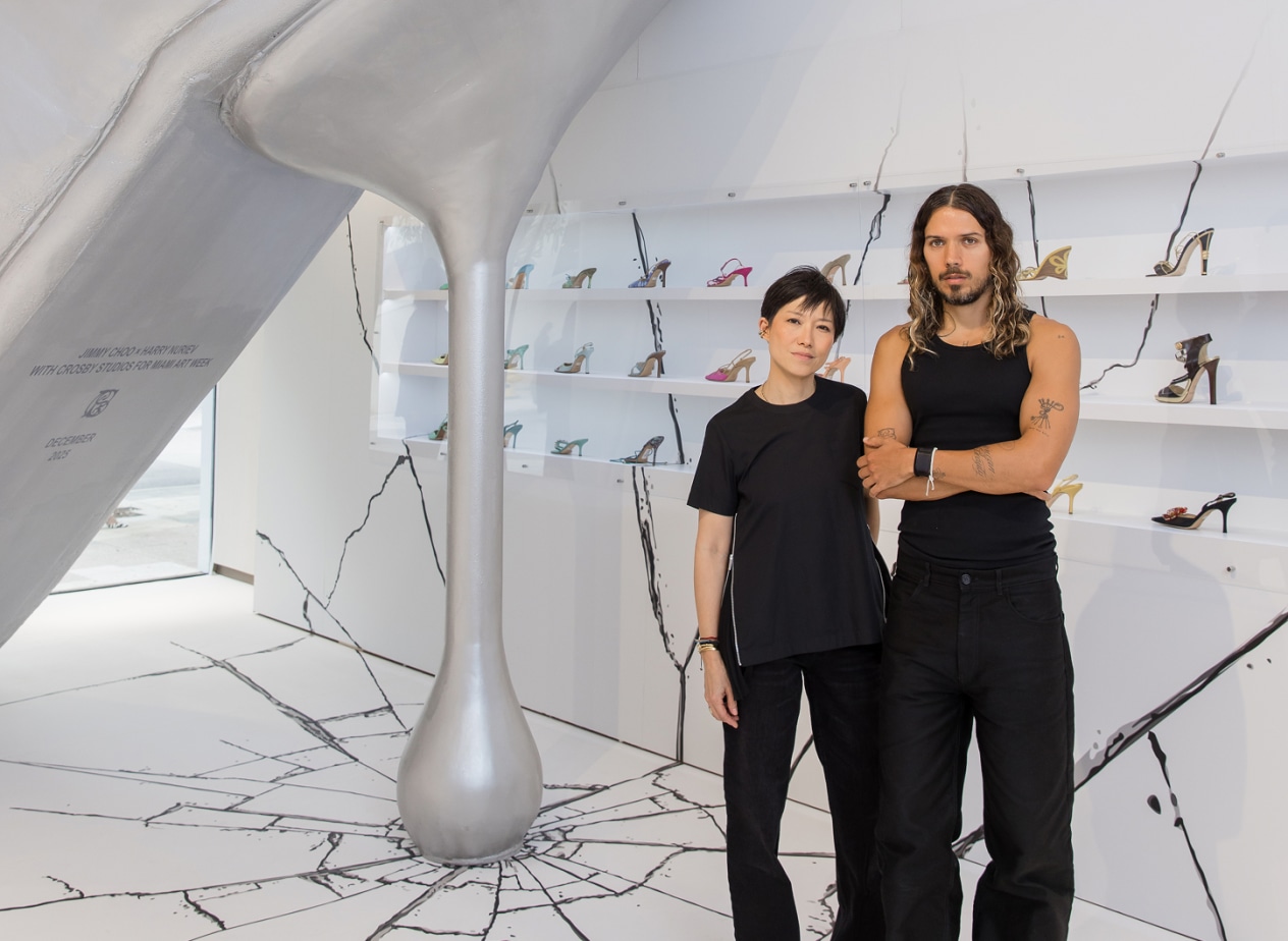  Interior view of the Jimmy Choo installation, showing the oversized silver high-heel sculpture and cracked floor design, with Sandra Choi and Harry Nuriev standing in front of wall-mounted shelves of shoes.