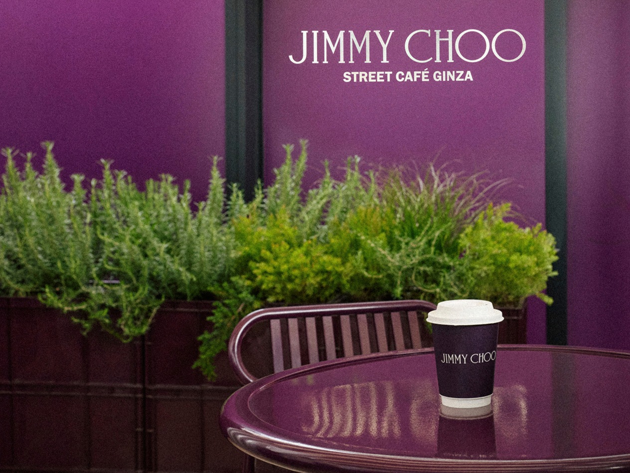 Coffee cup on a table outside Jimmy Choo Street Cafe Ginza with greenery and purple signage.
