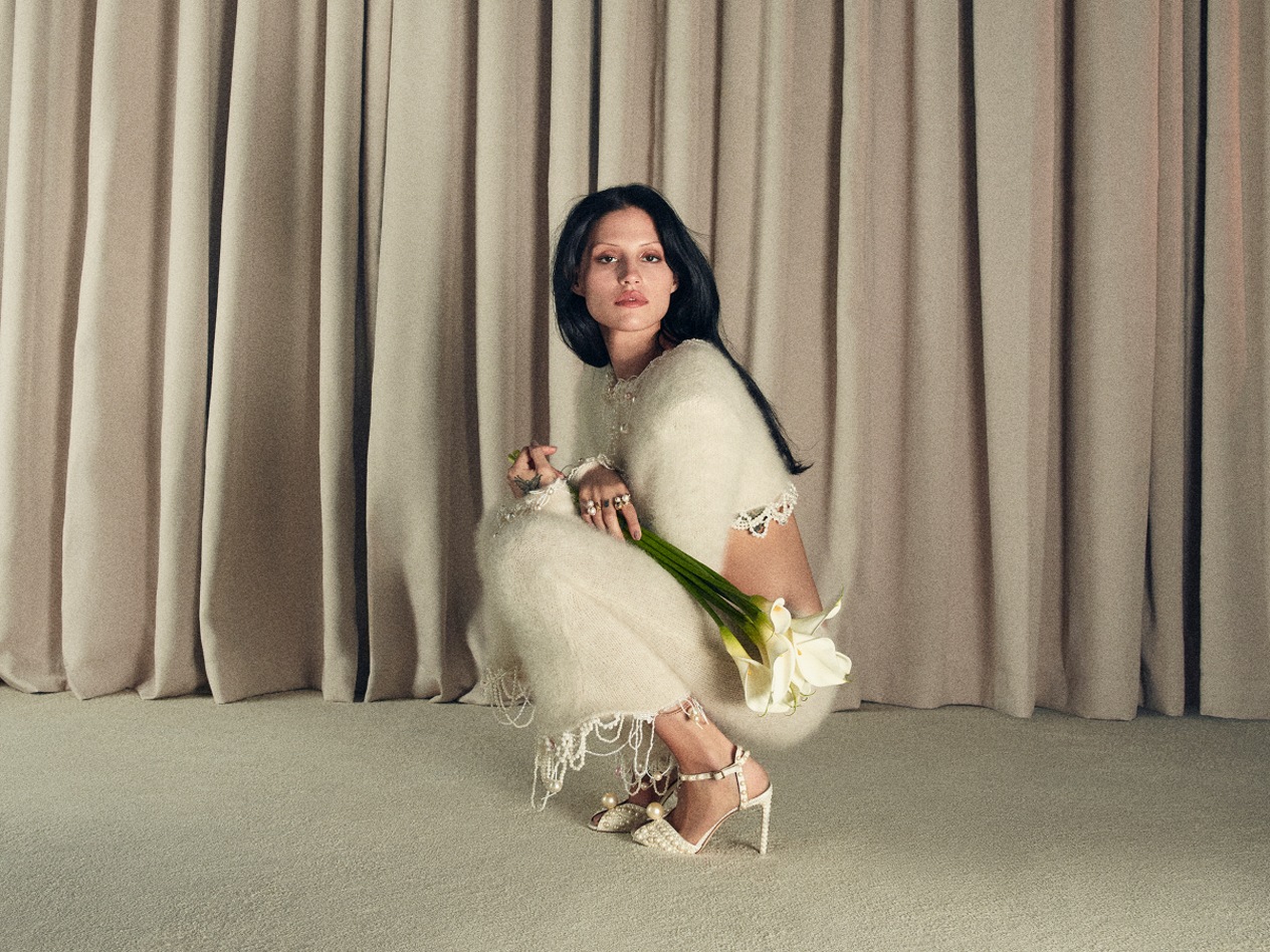Model crouching in ivory dress wearing open-toe bridal heels and holding white flowers.	