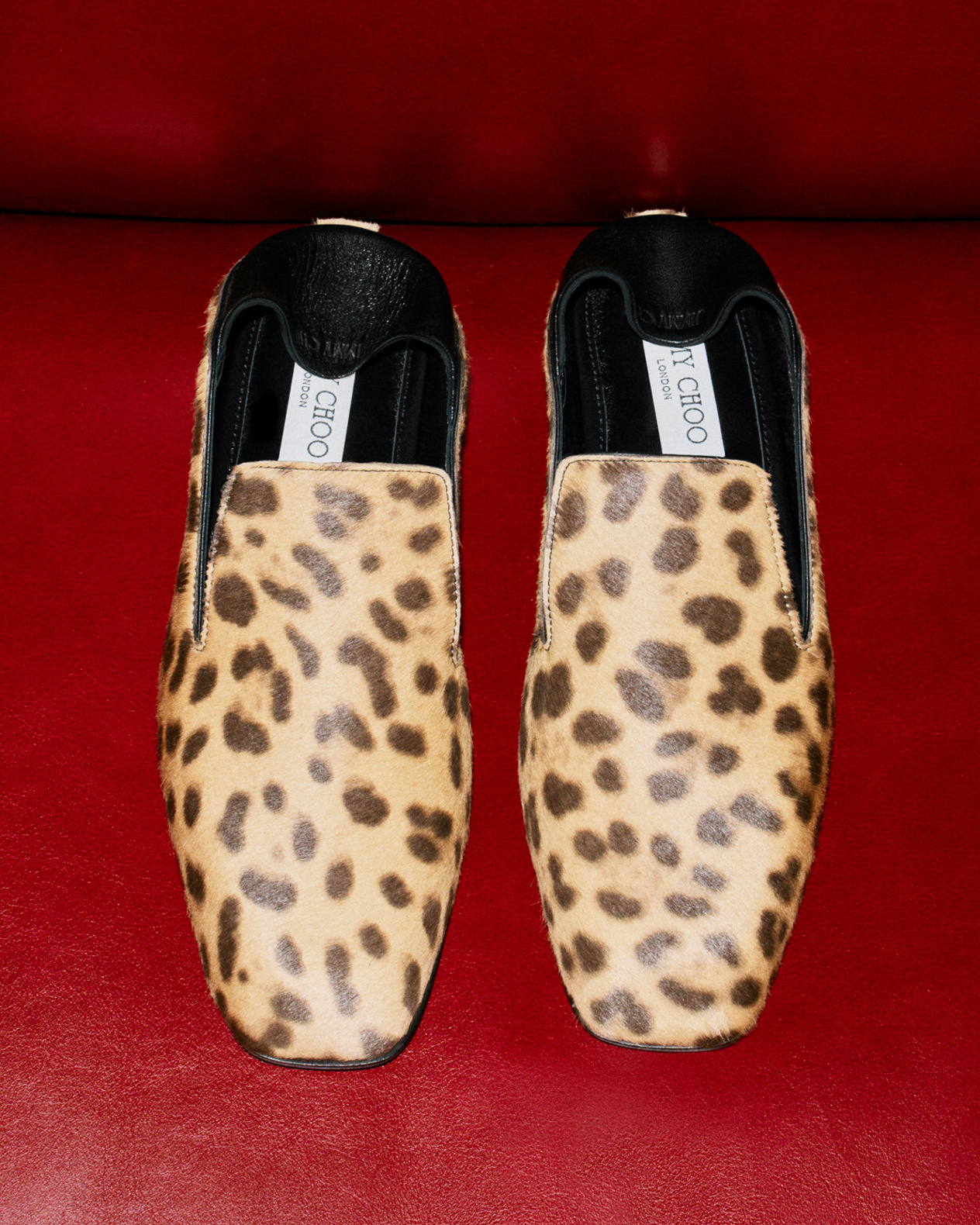 Leopard-print slip-on slippers with square toes and black leather trim, displayed against a bold red leather backdrop.