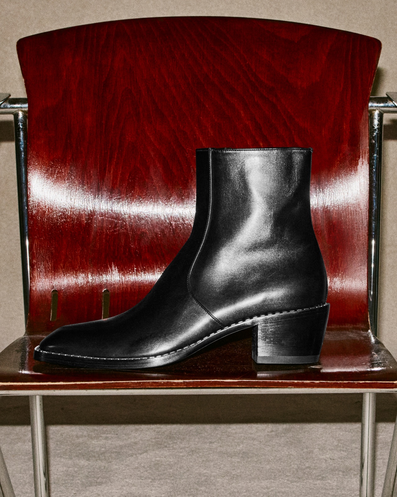 Black leather ankle boot with a mid block heel and sleek silhouette, displayed on a polished wooden chair with chrome details.