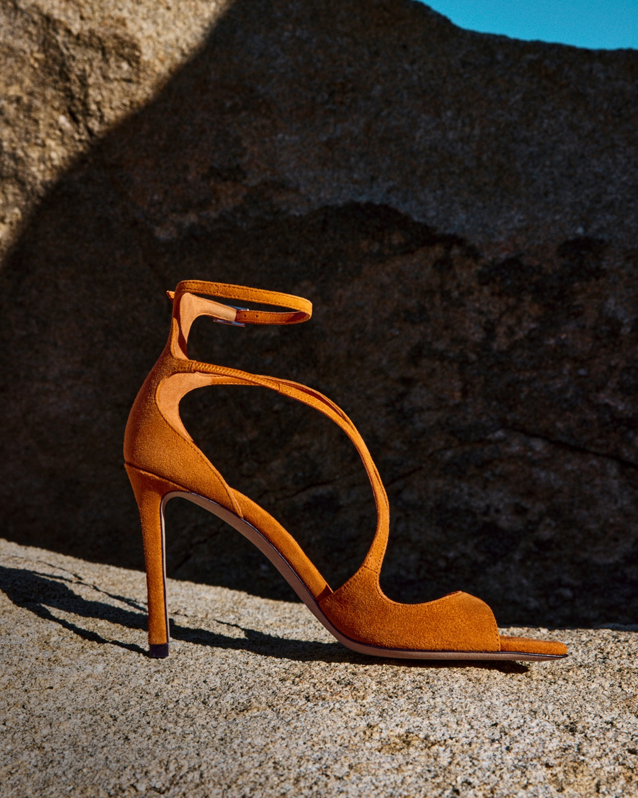 Orange suede heeled sandal with wraparound ankle strap displayed on rocky terrain.
