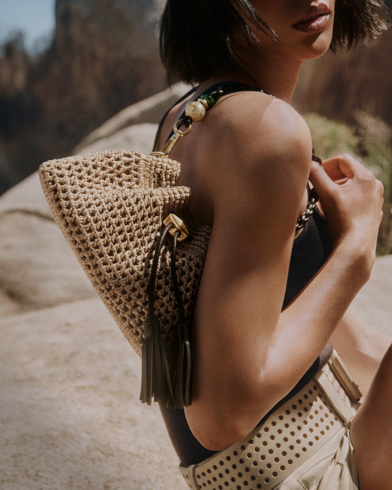 Woman carrying a woven raffia shoulder bag with tassel and bead detailing in a sunlit outdoor setting.