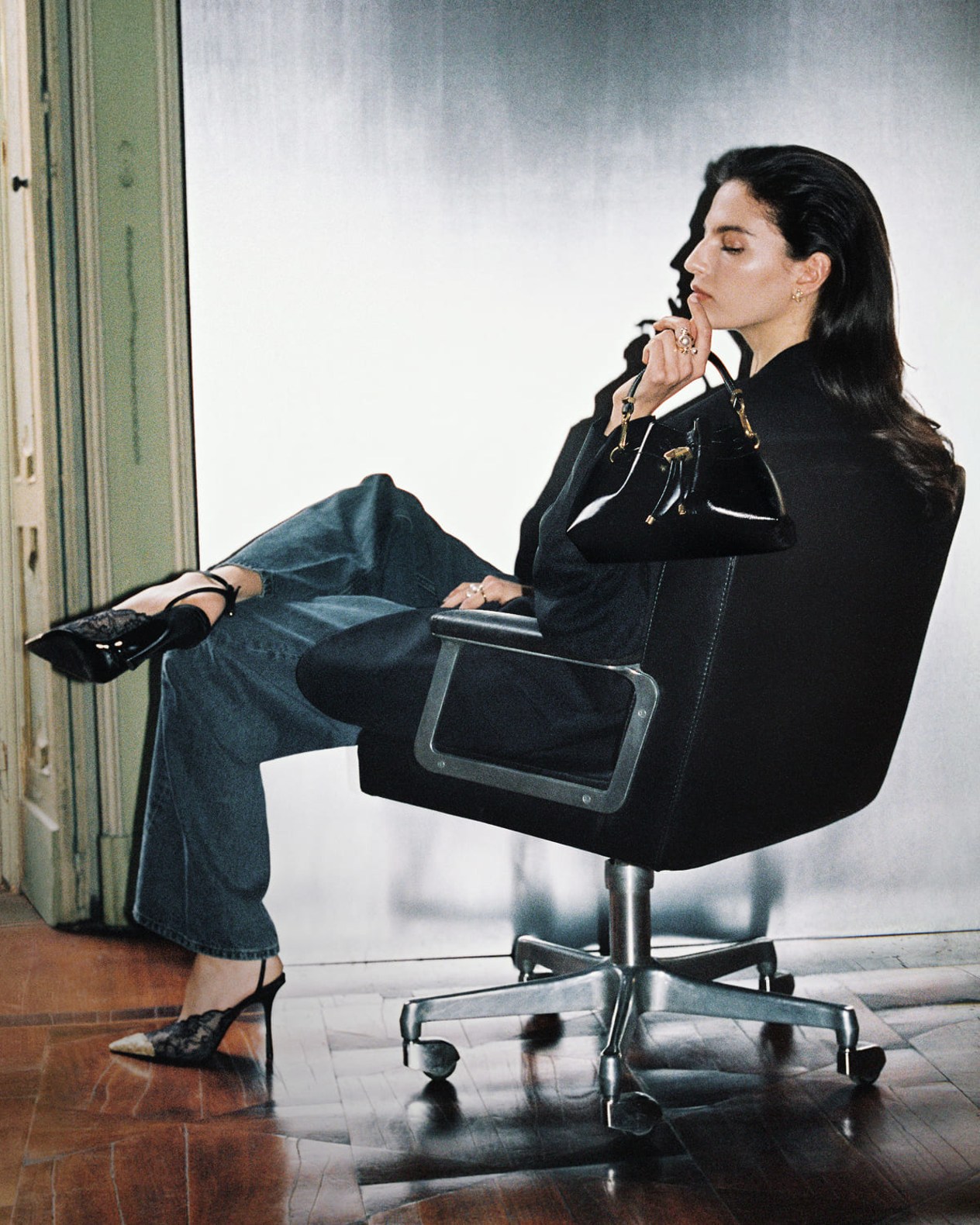 Model seated in an office chair wearing black lace pumps with metal lace toe-caps, holding a black leather top-handle bag.