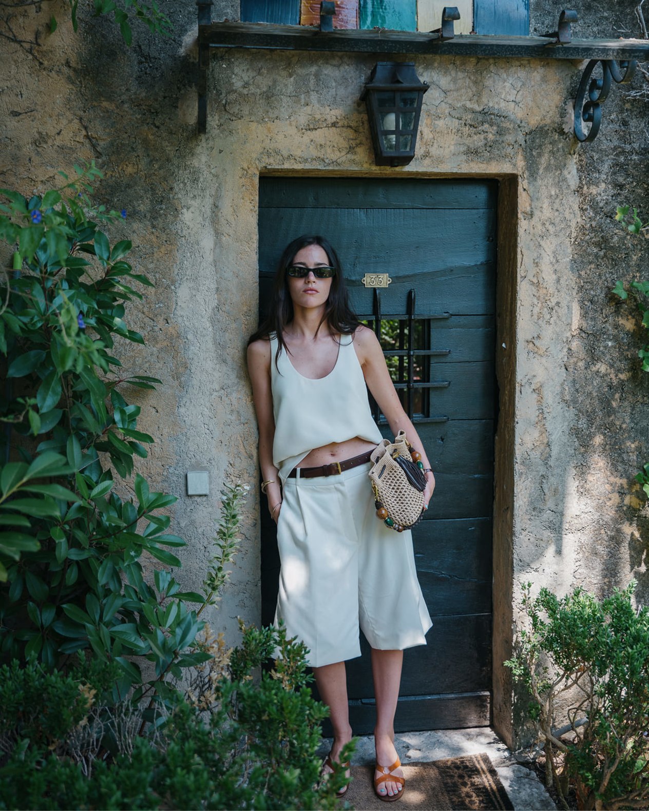 Model wearing a white sleeveless top, cropped trousers, and orange jelly sandals, carrying a woven drawstring bag while standing in a stone doorway.