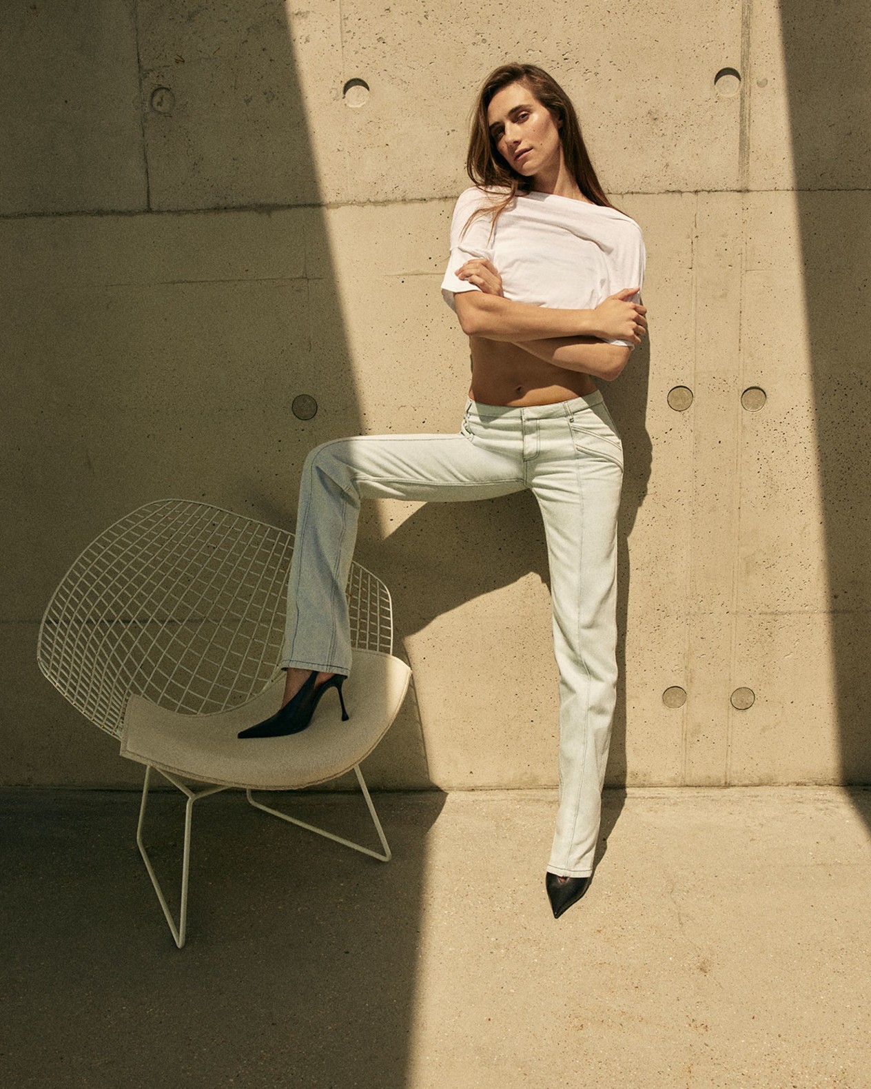 Devon Lee Carlson wearing a white T-shirt, light-wash jeans and black pointed pumps, posing against a concrete wall.