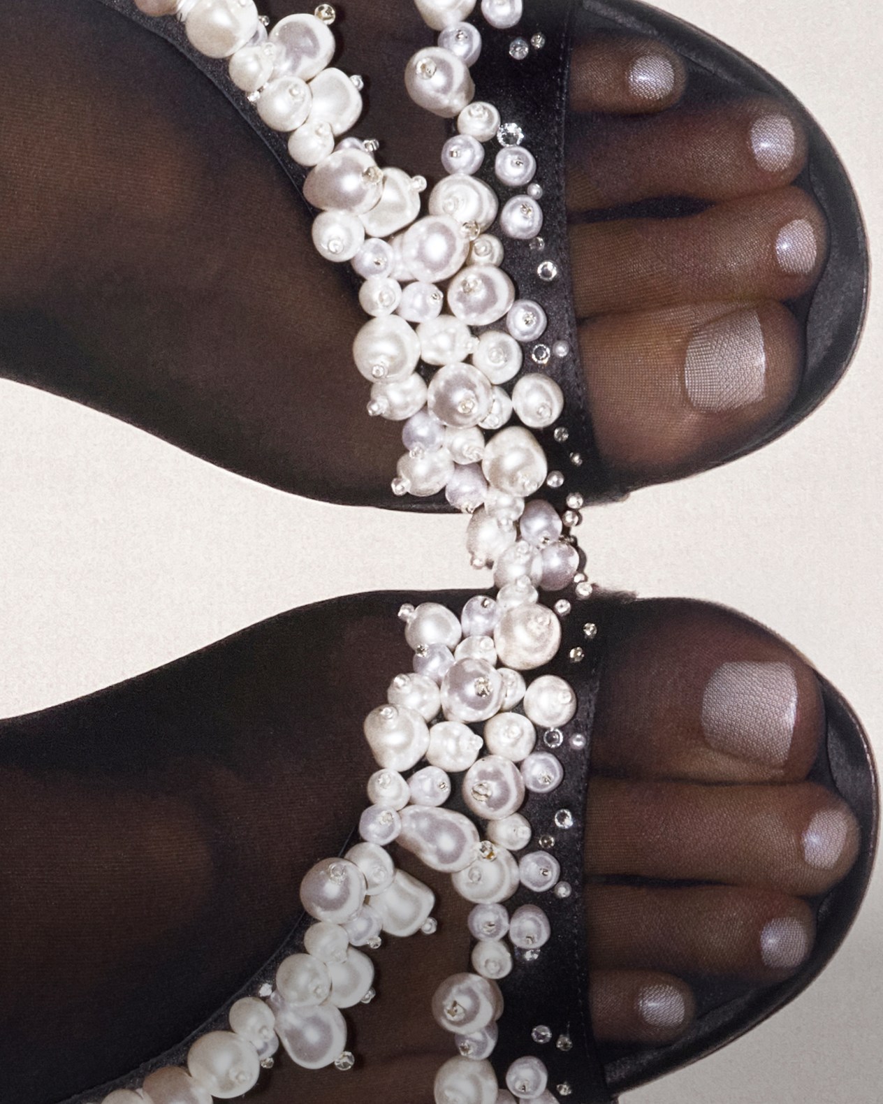 Close-up of black slingback sandals with stiletto heels and pearl and bead-embellished straps, worn by a seated model.