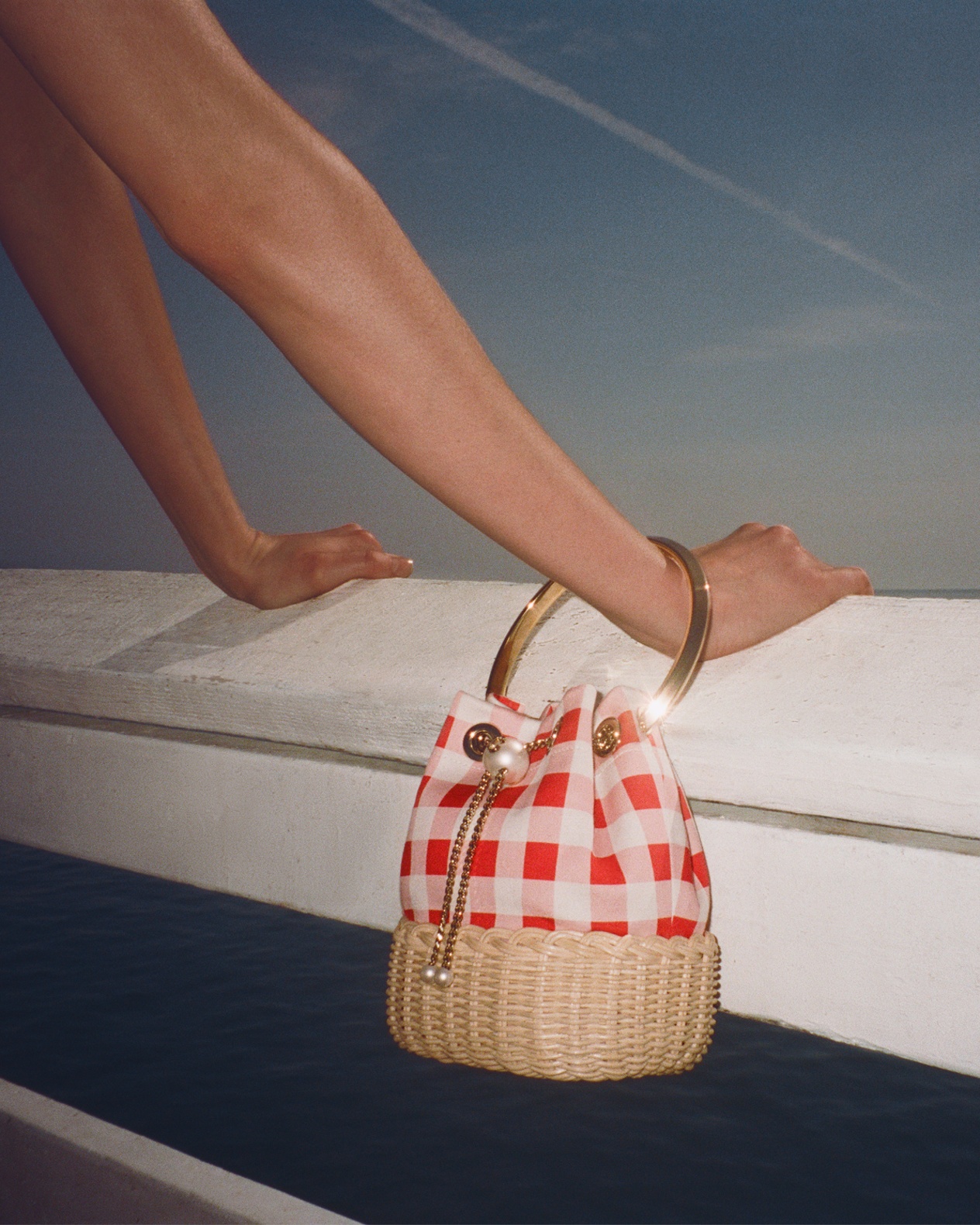 Red gingham and raffia bucket bag with gold bracelet handle resting on a stone ledge.