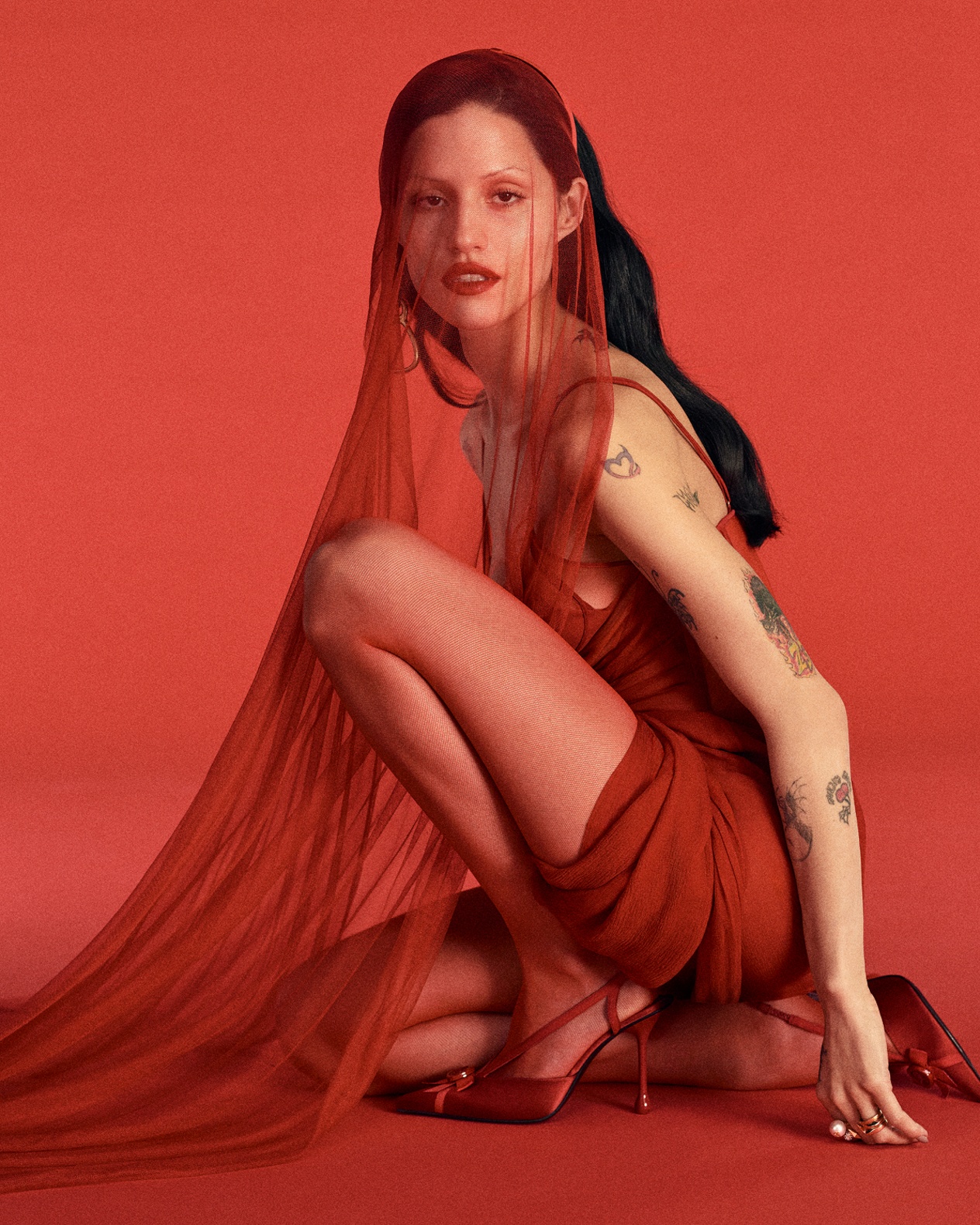 Model kneeling on a red backdrop wearing a red dress with sheer veil and matching red satin slingback heels. bag