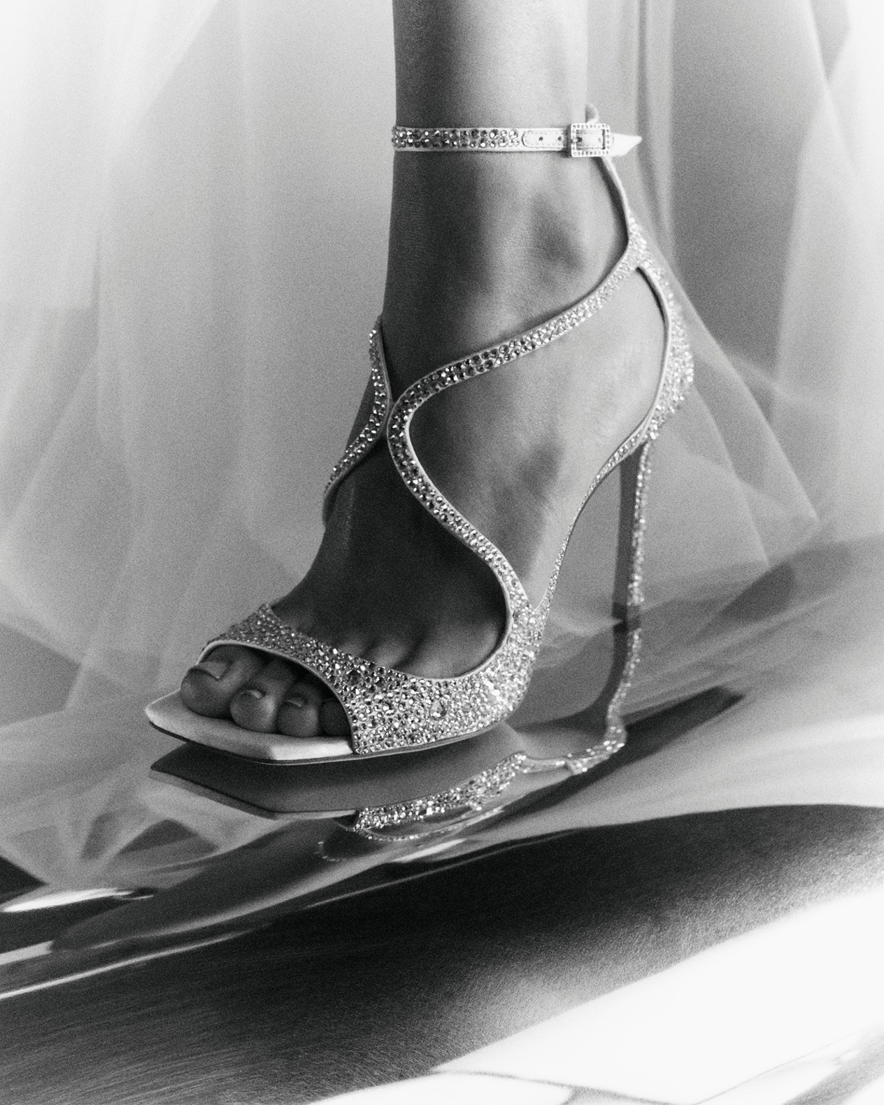 Black-and-white close-up of crystal-embellished heeled sandals with wraparound straps and mirrored reflection.