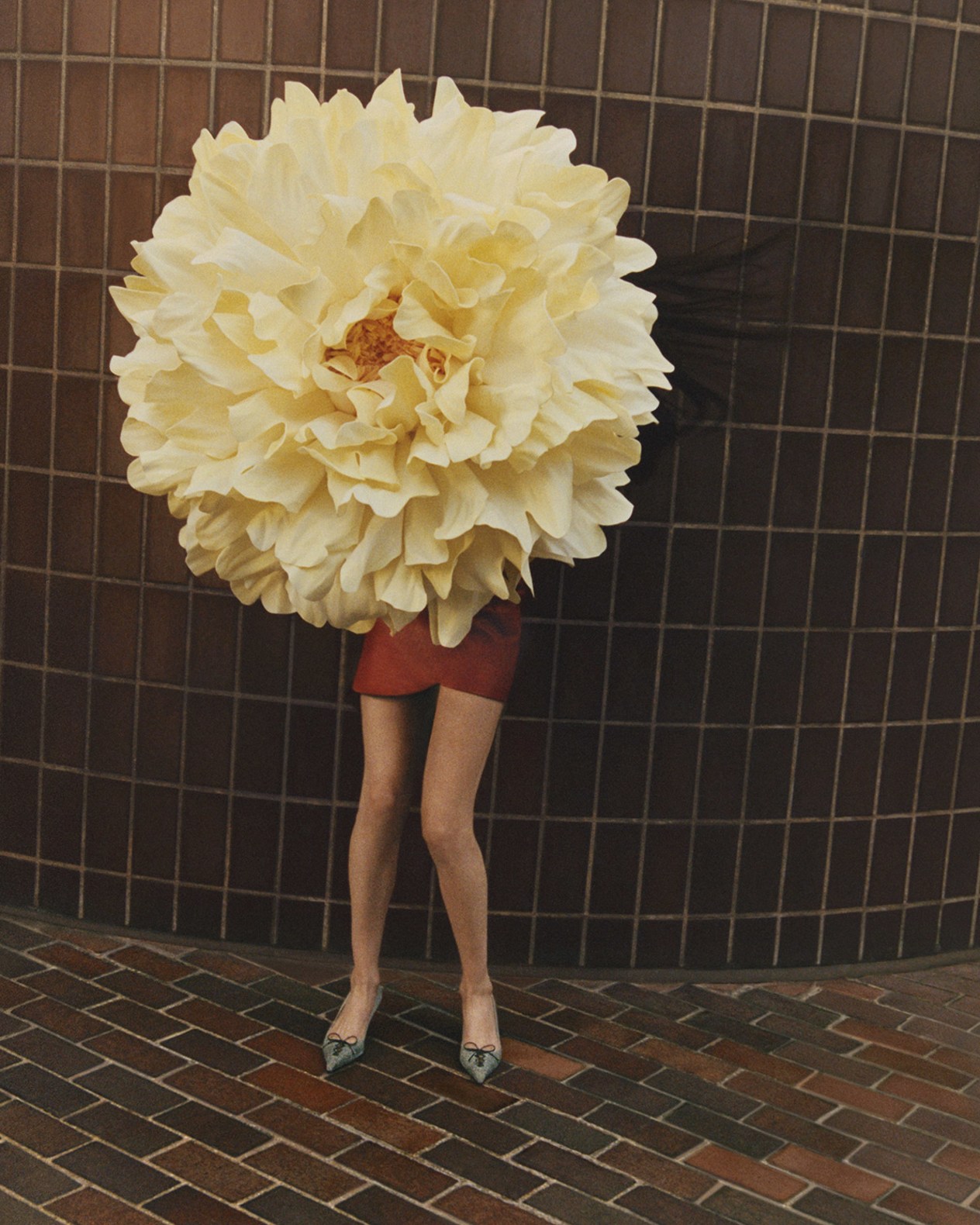 Woman standing against a tiled wall wearing a red dress and heels, with her upper body completely obscured by an oversized pale yellow flower positioned in front of her face and torso.