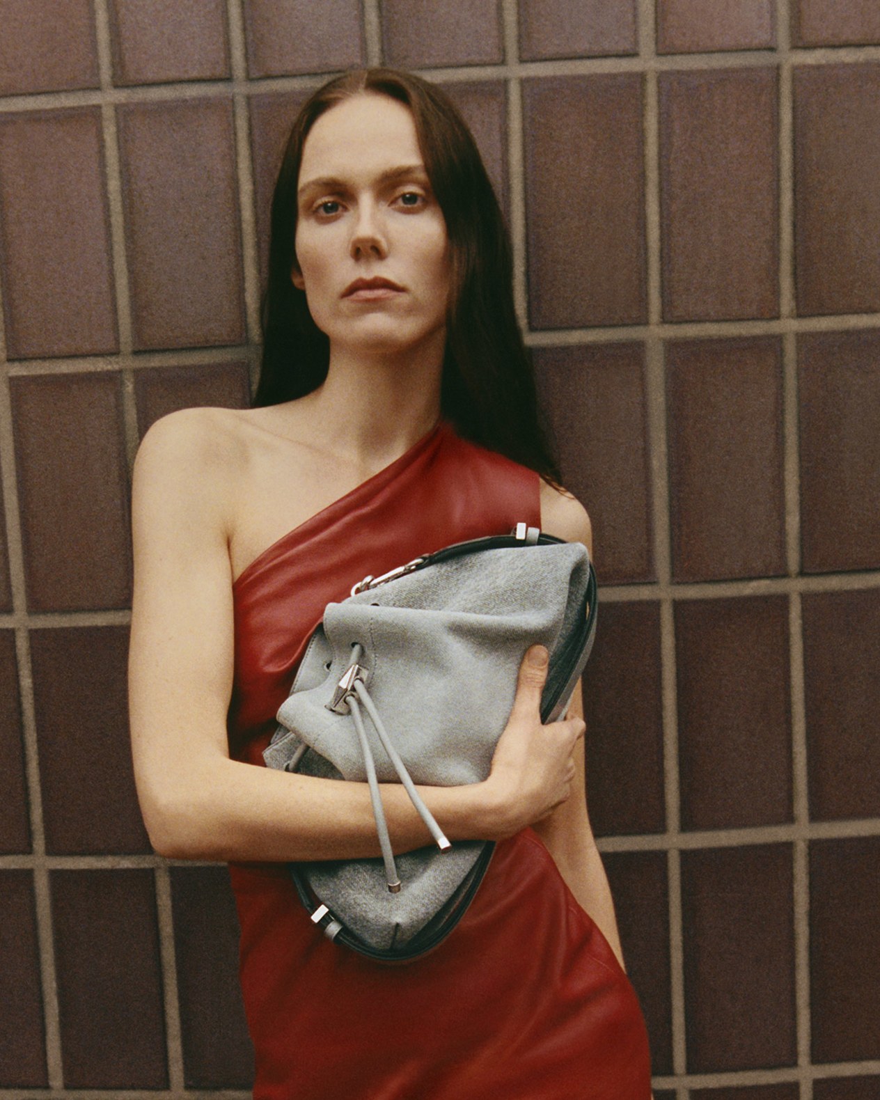Woman in a one-shoulder red dress holding a light grey drawstring bag across her chest, standing against a brown tiled wall.