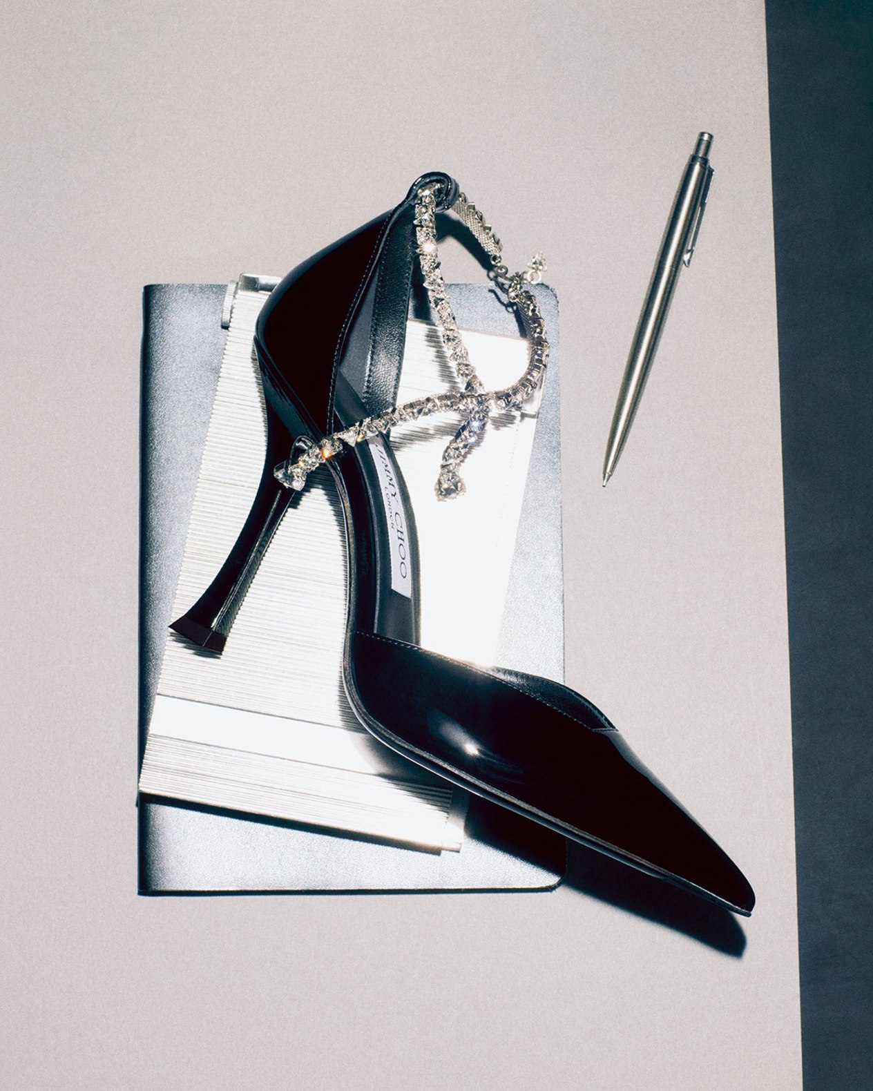 Black patent pump with a crystal-embellished ankle strap, styled on a white book with a metallic pen beside it.