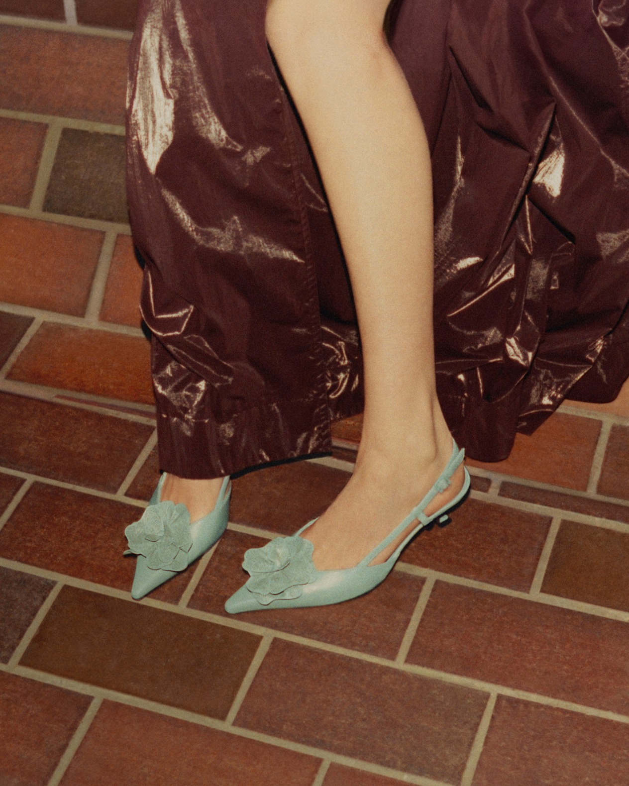 Close-up of a woman&rsquo;s legs wearing pale green pointed slingback heels with floral detailing, standing on a brick floor.	