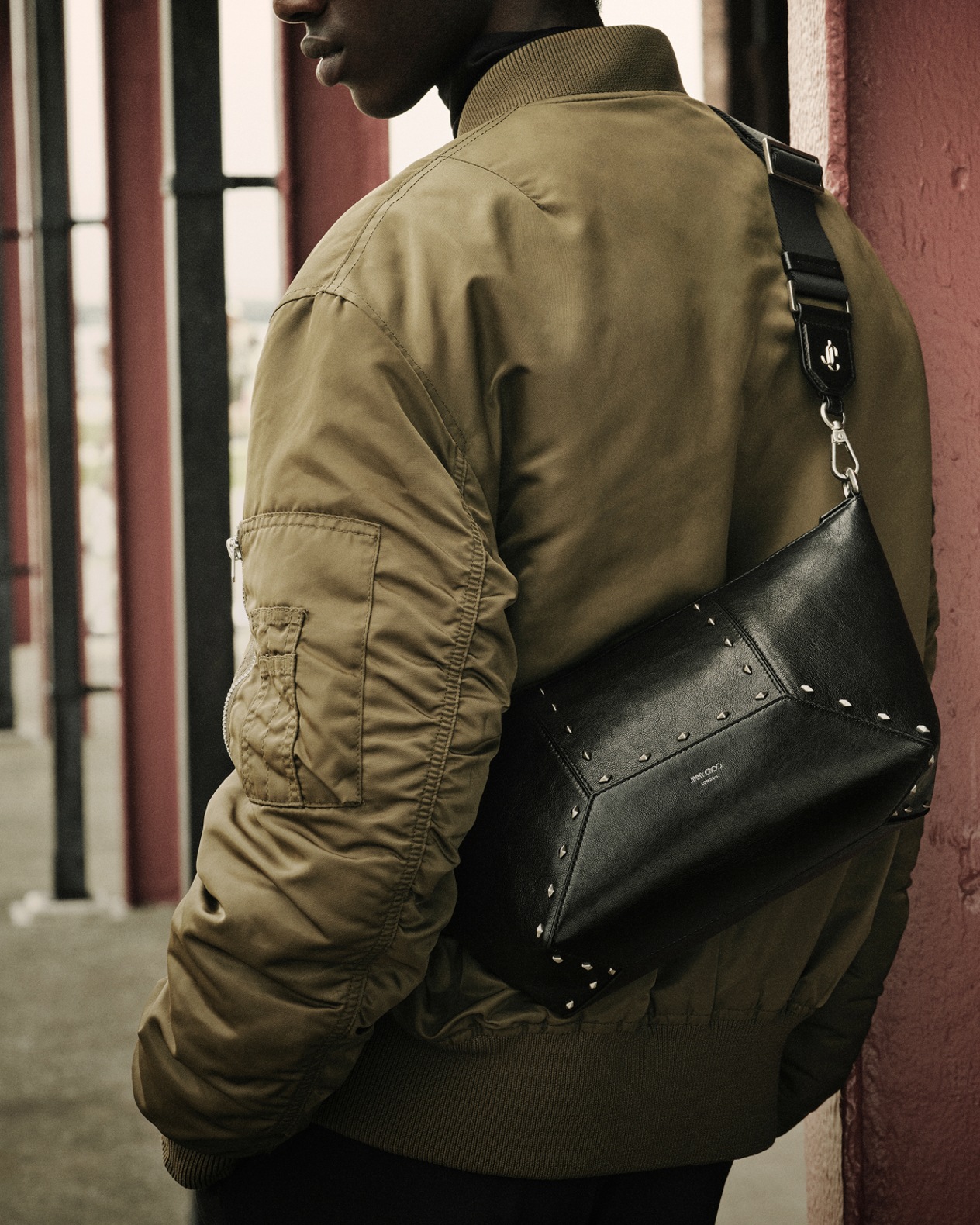 Male model in khaki bomber jacket  wearing a crossbody black leather bag with oversized buckle detail.