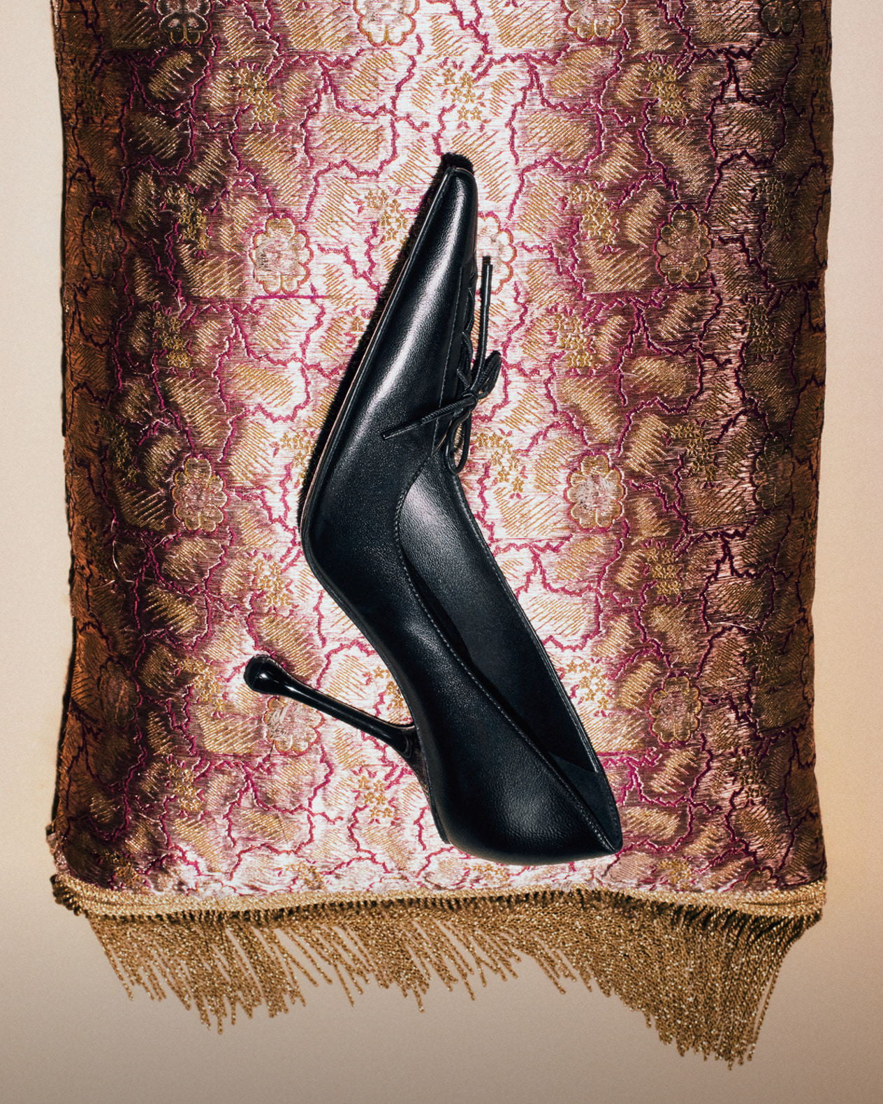 Black leather pointed-pump with a sculptural drop heel, displayed against a textured gold and burgundy fabric backdrop.