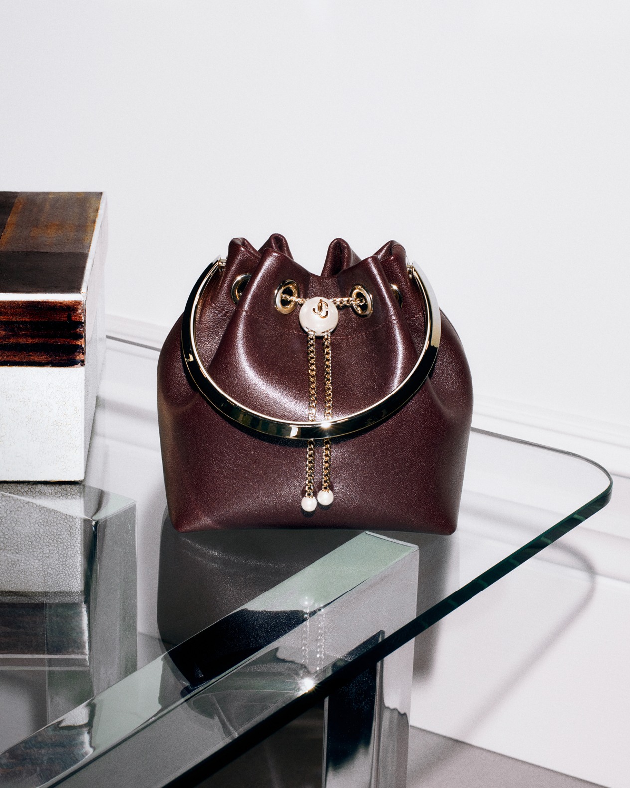 IMAGE ALT: Merlot leather drawstring bag with a gold chain and pearl detail, styled on a glass table.