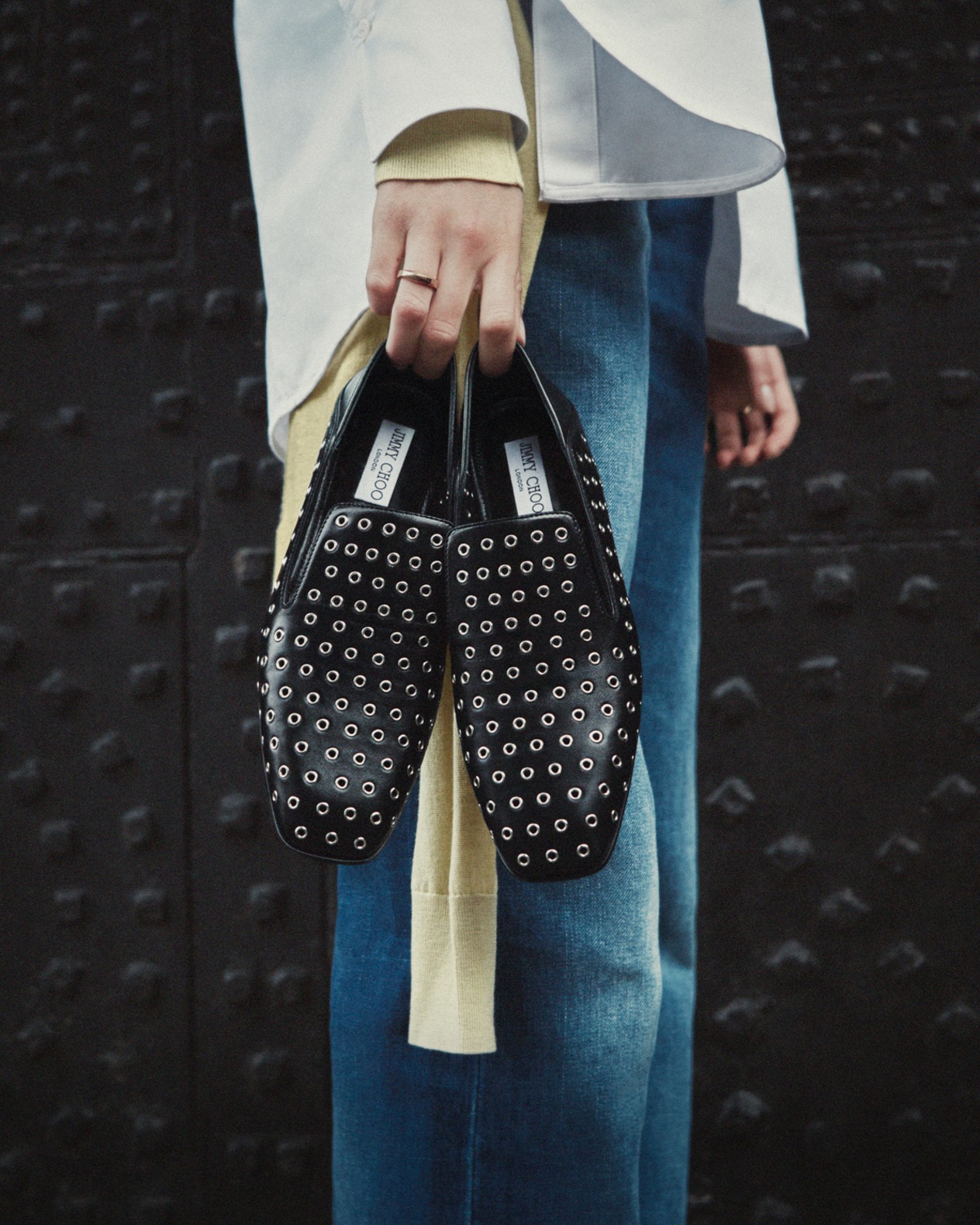 Person holding a pair of black studded Eliot slippers by the heels, styled with blue jeans and a light shirt, photographed against a dark textured background.