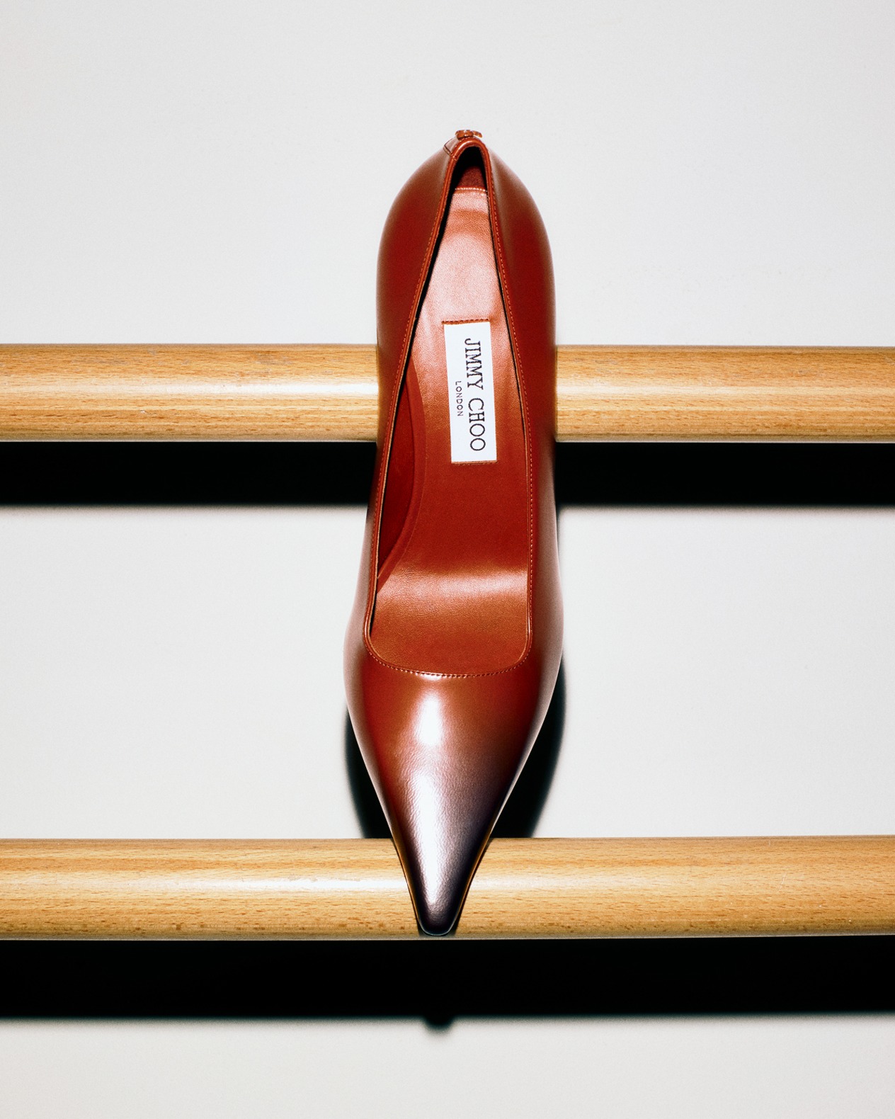 Single brown leather pointed-toe pump with an ombre gradient effect, balanced on a wooden ballet barre against a white background.