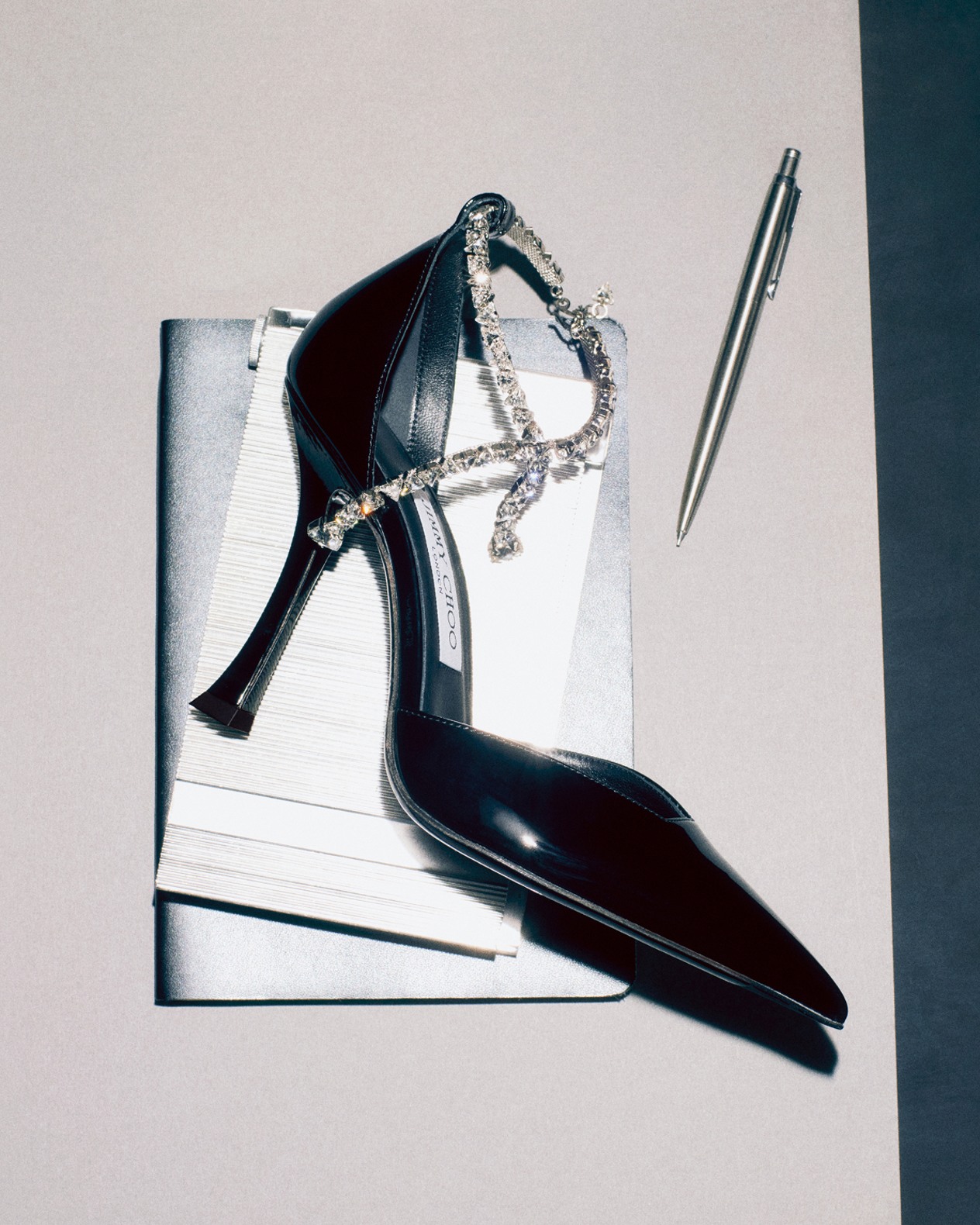 Black patent pump with a crystal-embellished ankle strap, styled on a white book with a metallic pen beside it.