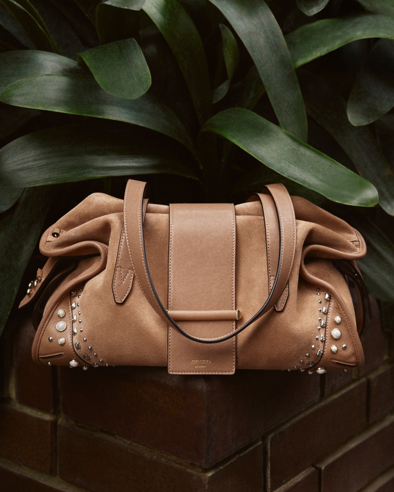 Light brown top-handle handbag with structured silhouette placed on a brick floor against a textured stone wall.