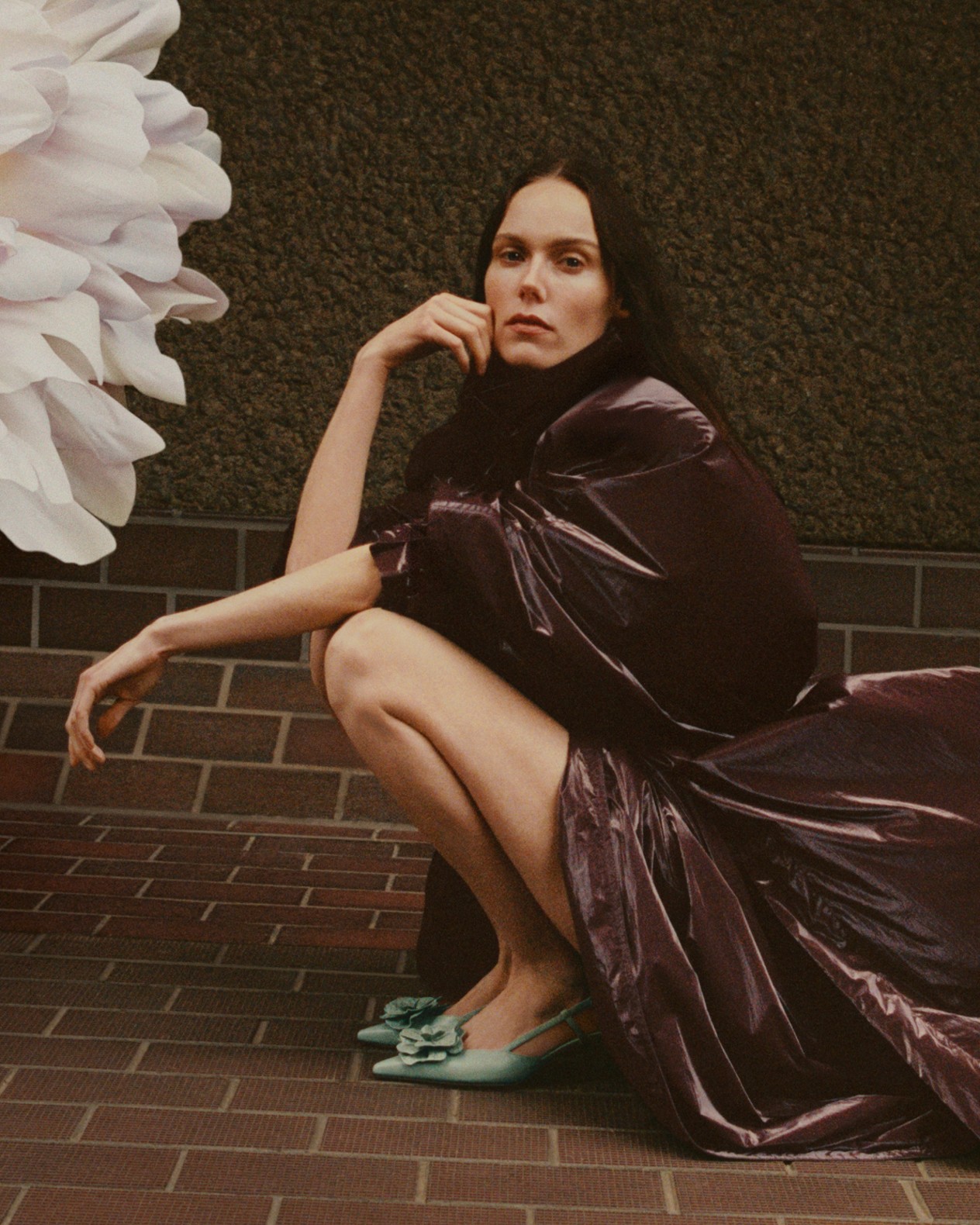 Woman crouching on a brick pavement wearing a dark, glossy dress and pale green pointed slingback heels with floral corsage accent, against a textured wall.
