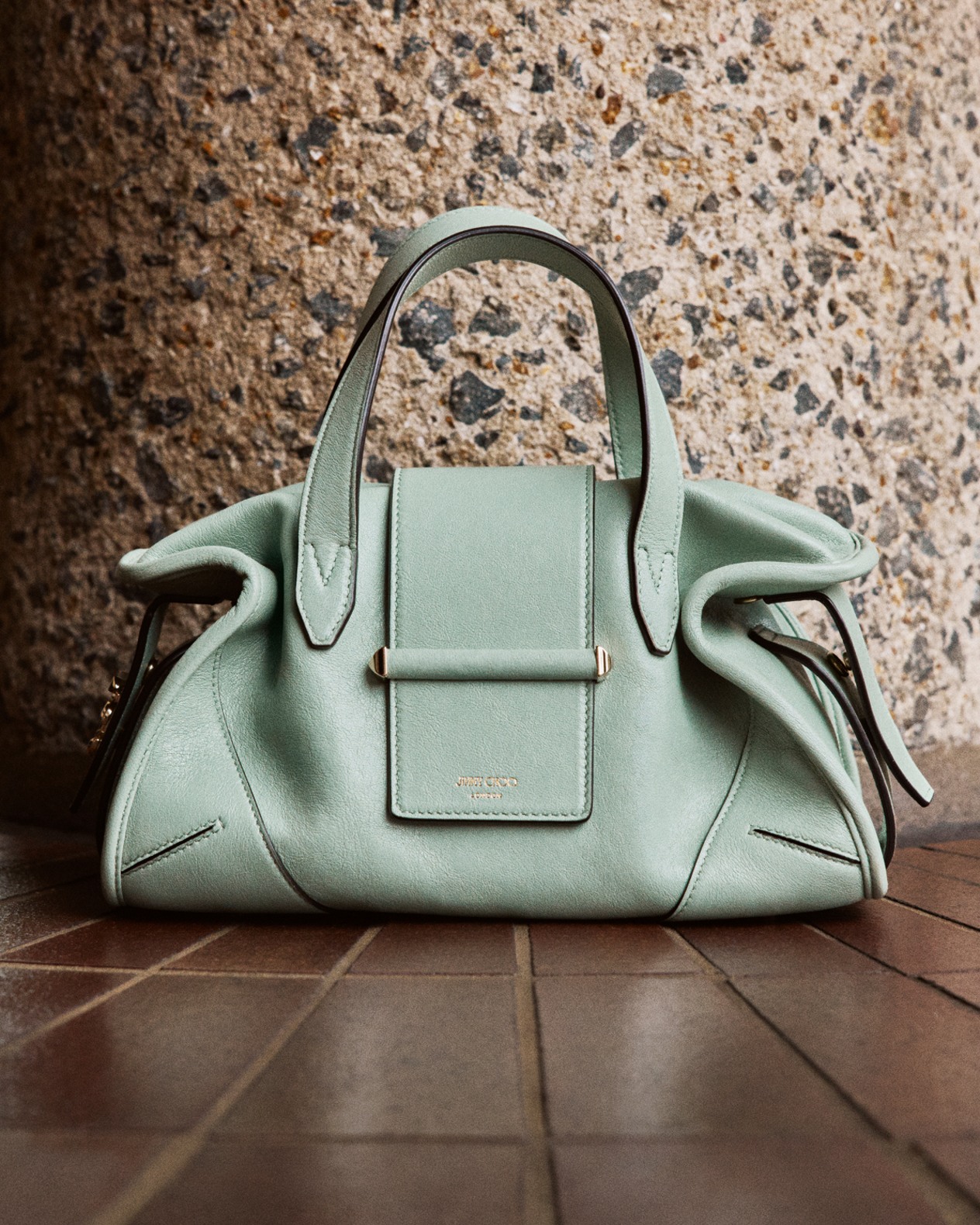Mint green top-handle handbag with structured silhouette placed on a brick floor against a textured stone wall.	