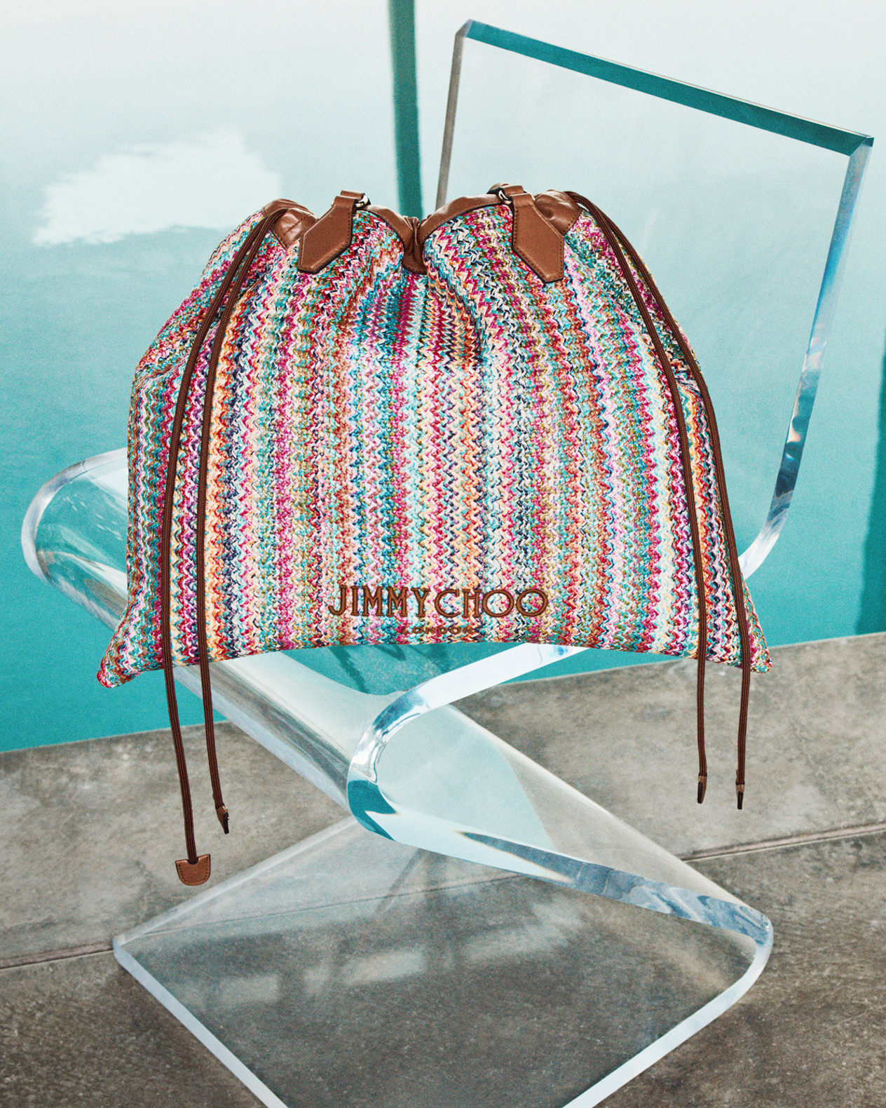 Multicolour woven drawstring bag with leather ties placed on a clear chair beside a pool.