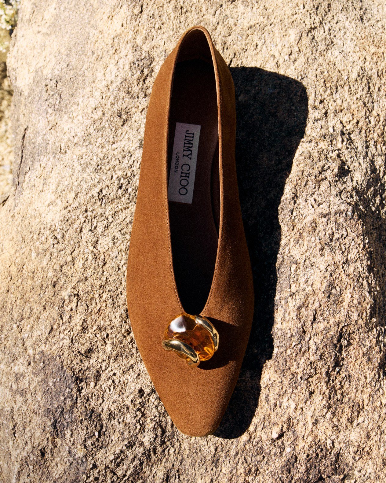 Beige flat sandals with textured fringe straps placed on rocky ground.