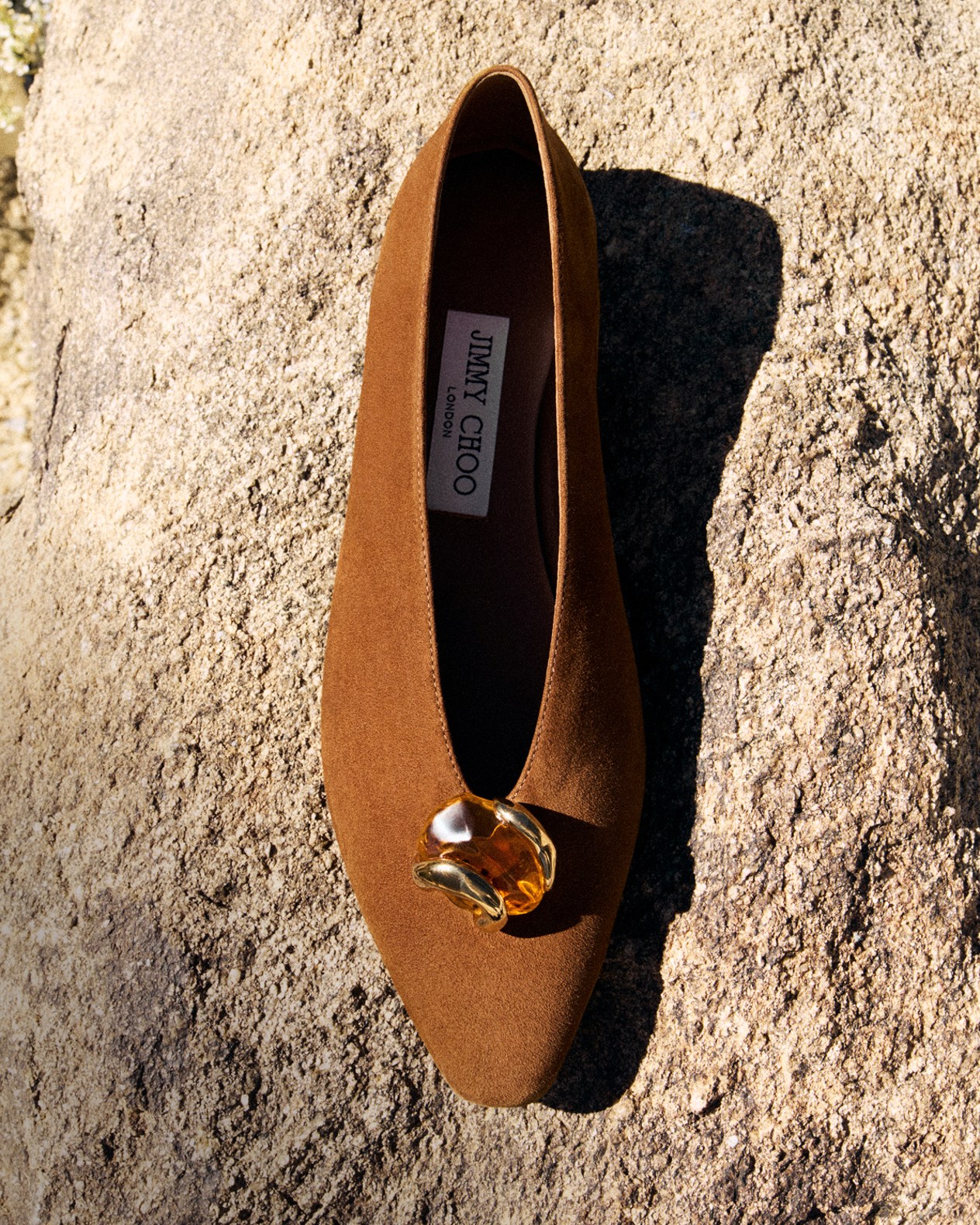 Beige flat sandals with textured fringe straps placed on rocky ground.