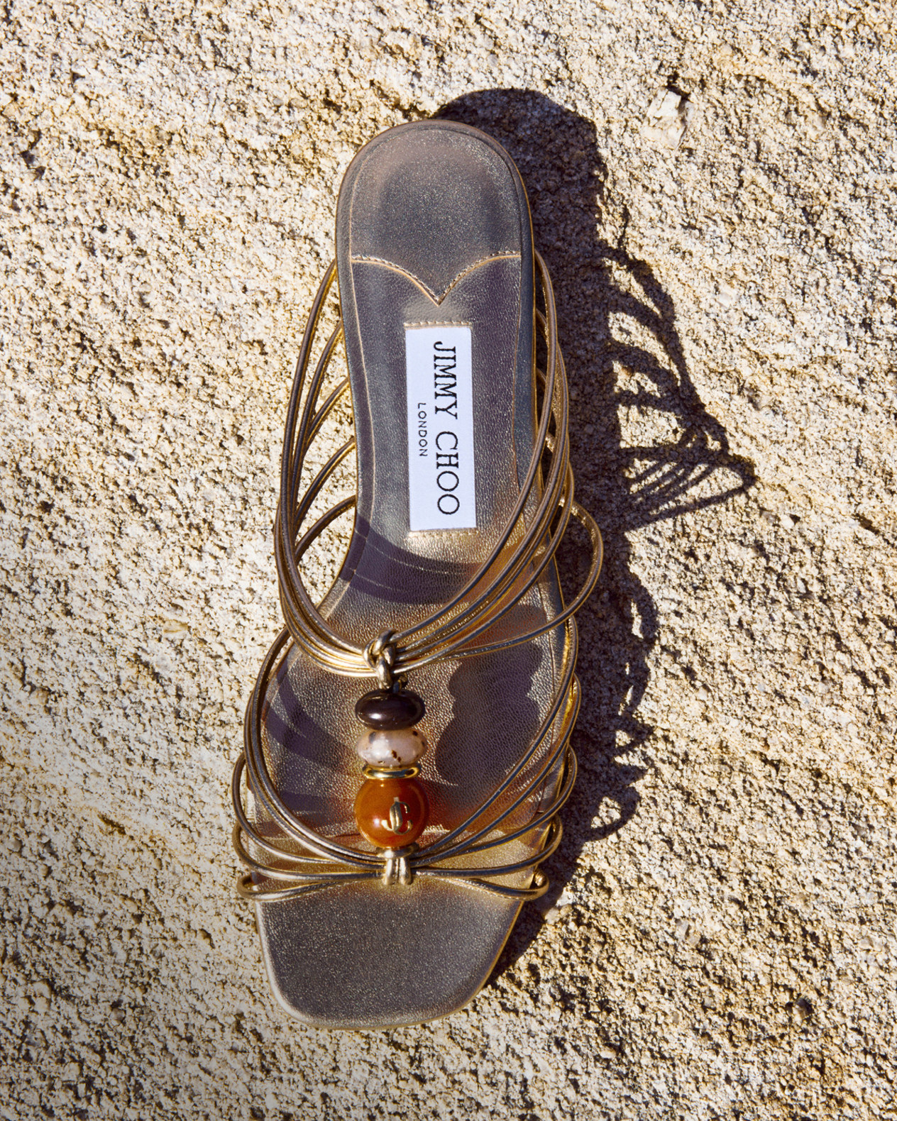 Brown suede flat shoe with a sculptural gold stone embellishment displayed on a textured stone surface.