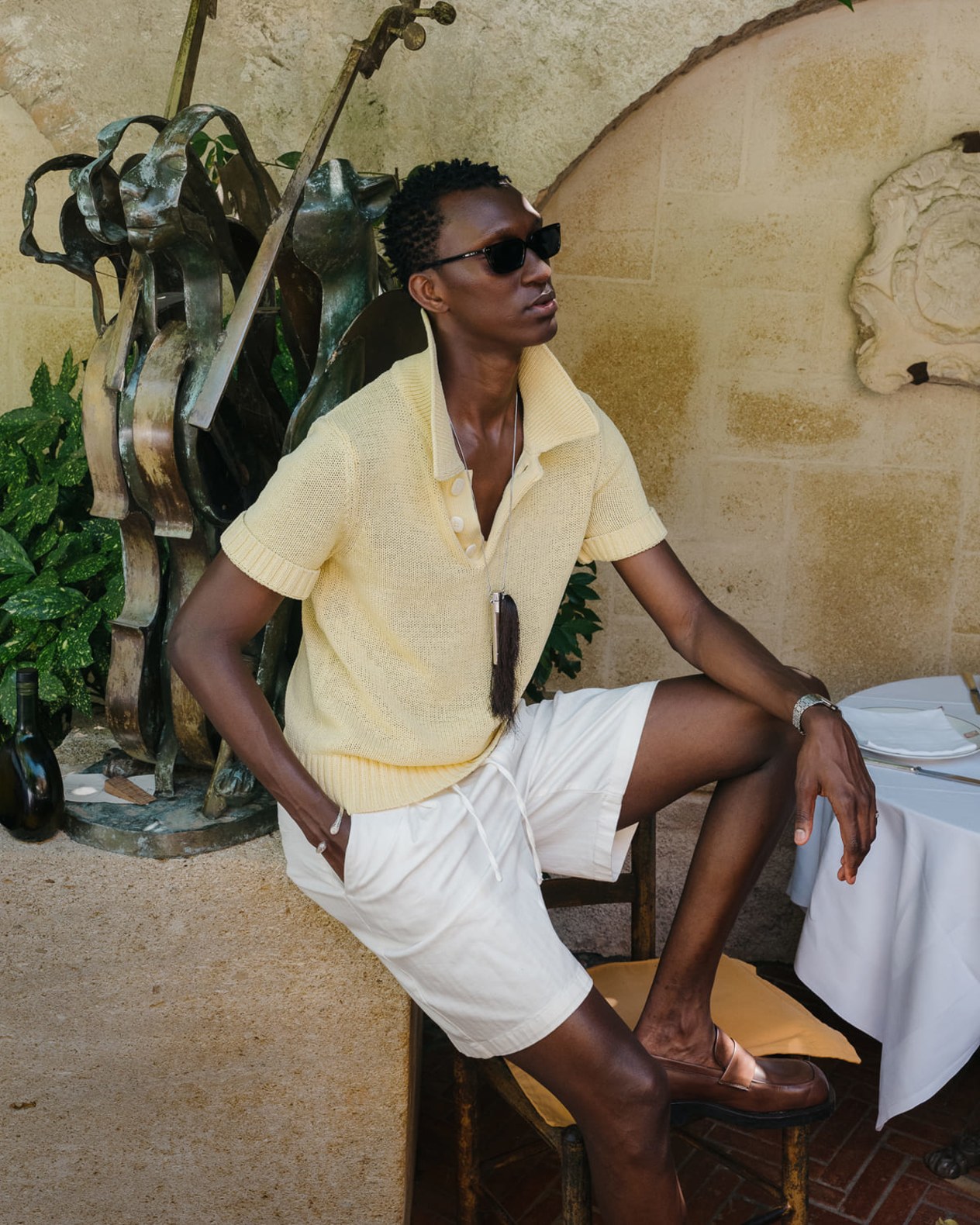 Model wearing a blue striped knit top, denim skirt and green wedge sandals, seated on a rustic sofa.
