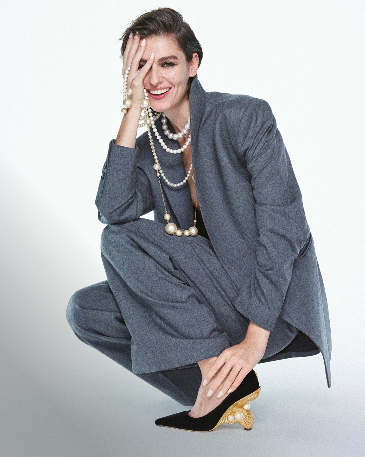 Model crouching in a grey oversized suit and layered pearl necklaces, wearing black pointed-toe pumps with sculptural gold heels and pearl detailing.