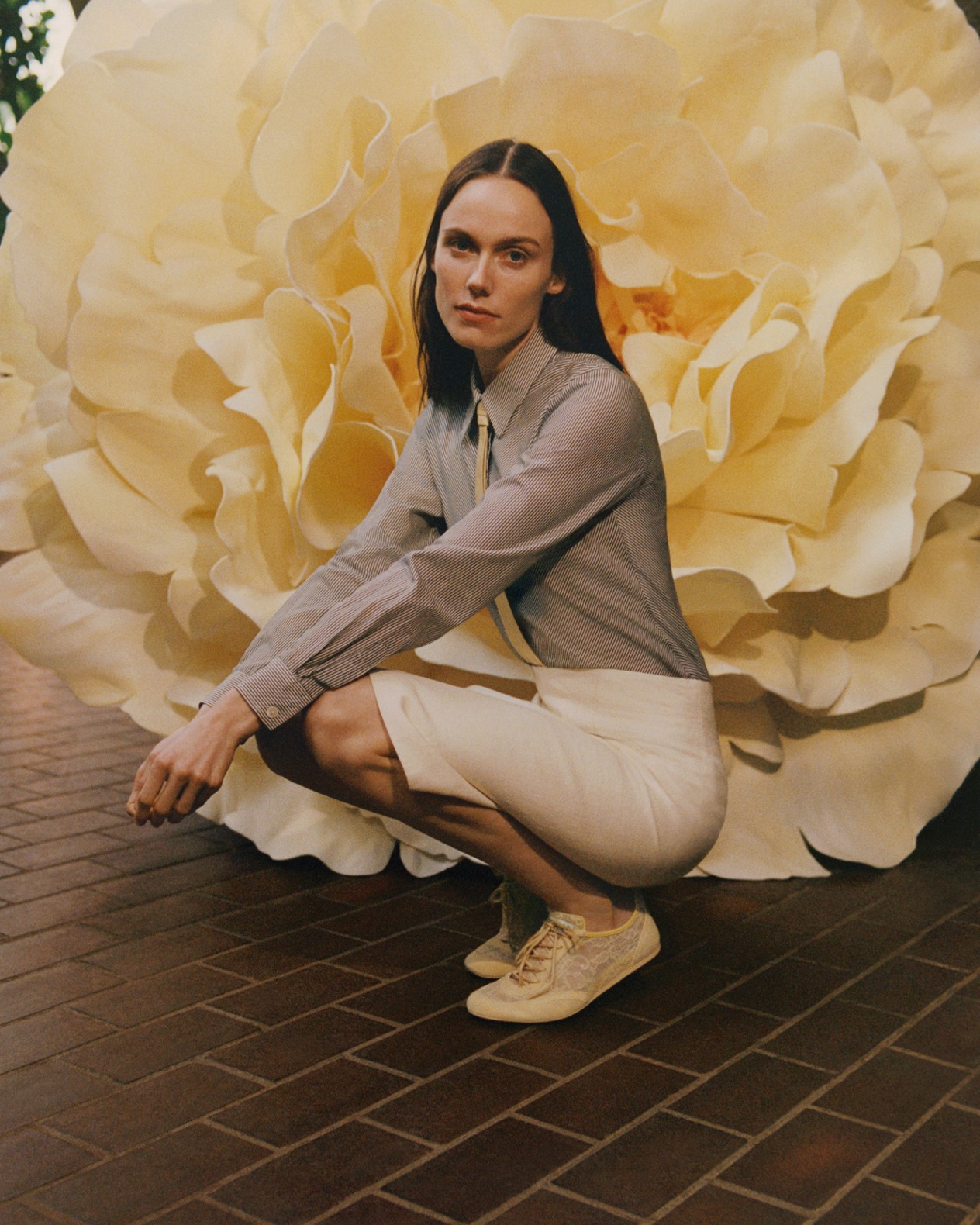 Model crouching outdoors wearing a light knit top, cream skirt and light yellow lace trainers, set against an oversized floral backdrop.